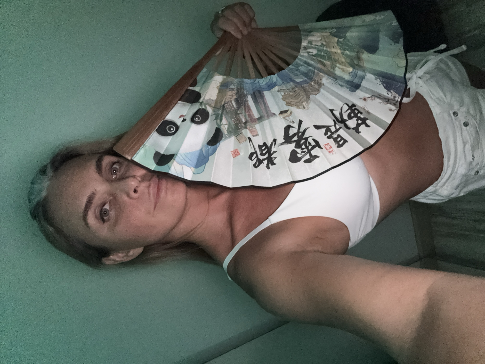 A blonde woman with graying hair and fair skin is holding a folding fan in front of her face, obscuring half of it. She is wearing a white bralette and white drawstring shorts. The fan has an image of a panda on it and some Asian characters. The background is a plain teal wall, and the lighting is dim and green-tinted. The image appears to be a selfie. The location is not discernible from the image.
