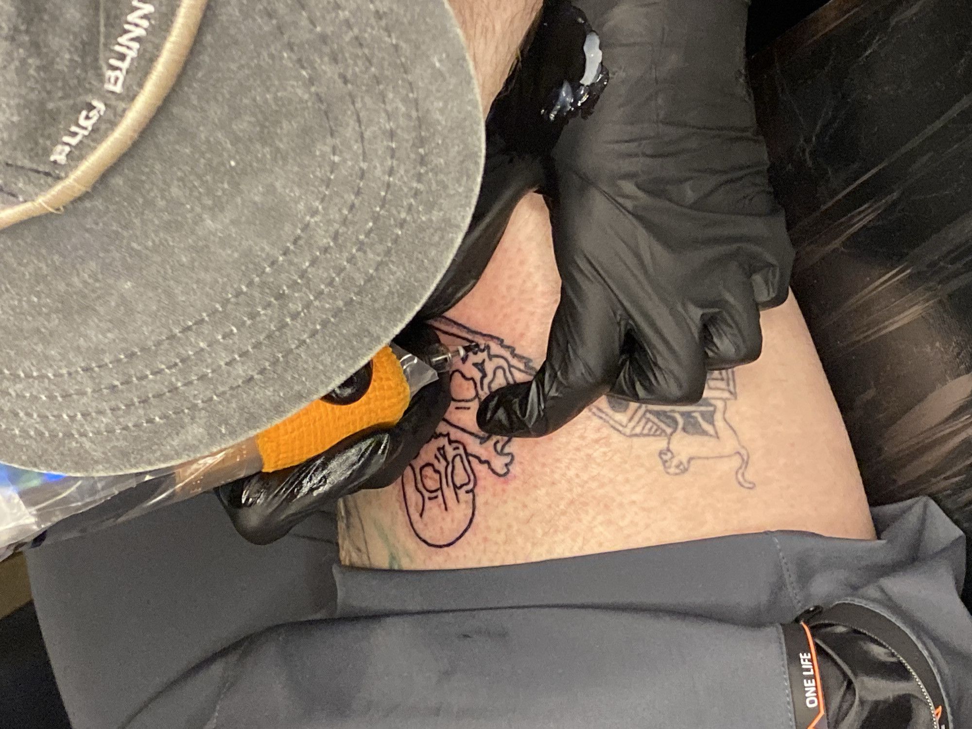 A person is getting a tattoo on their leg. The tattoo artist is wearing black gloves and a grey baseball cap. The person receiving the tattoo is wearing grey pants with a black accent and the words "ONE LIFE" visible on a tag. The tattoo being applied is a stylized skull and crossbones. Another tattoo, possibly depicting a lion or similar feline, is already present on the leg. The location is not discernible as Ushuaia, Argentina.