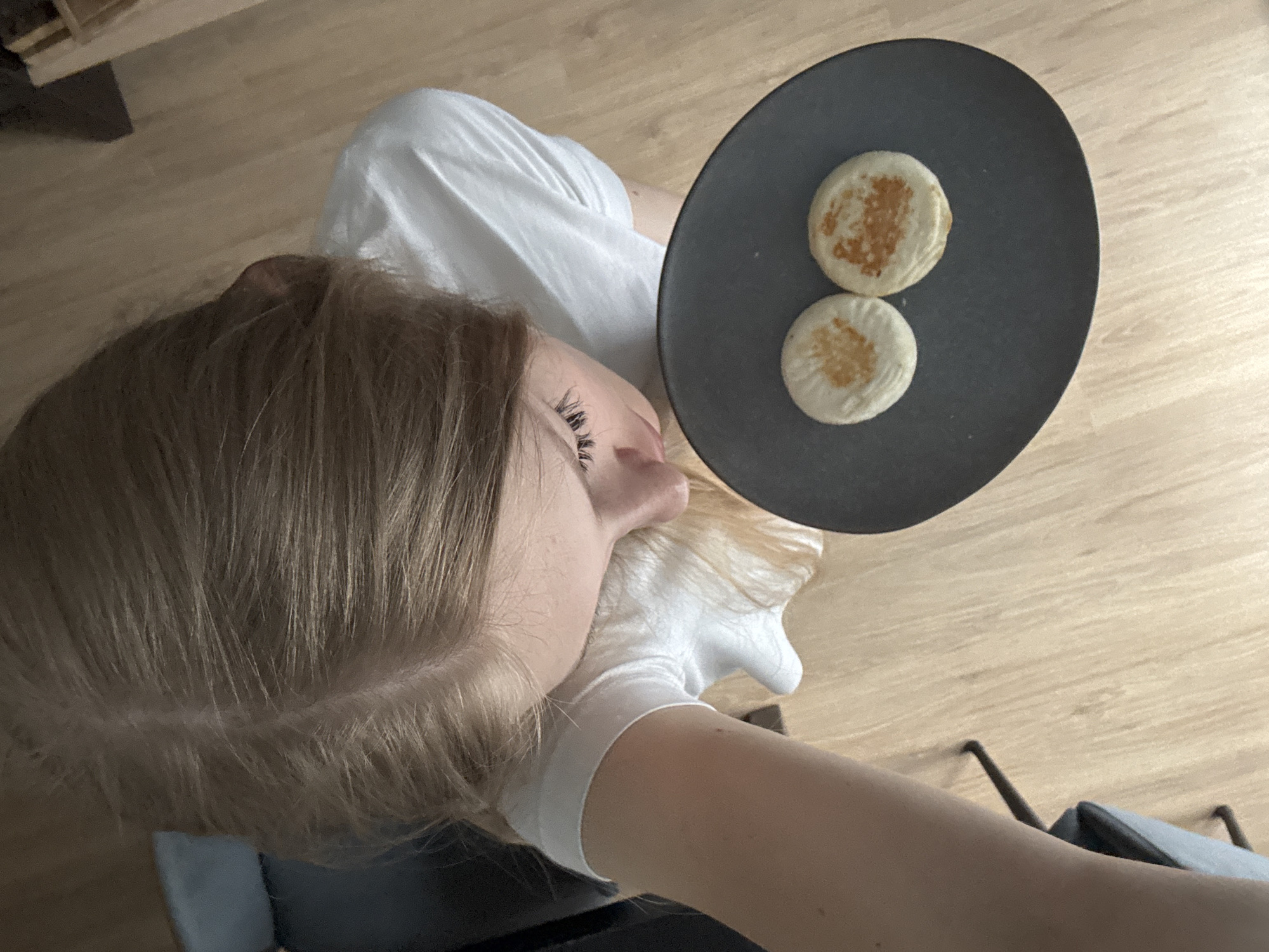 A young woman with blonde hair is looking down at a dark plate holding two round, golden-brown pastries. She is wearing a white t-shirt and is positioned to the left of the plate. The scene appears to be indoors, with a wooden floor visible. The lighting suggests it could be daytime. The woman's face is partially obscured as she leans forward. No specific location in the city of Perm, Russia can be determined from the image.