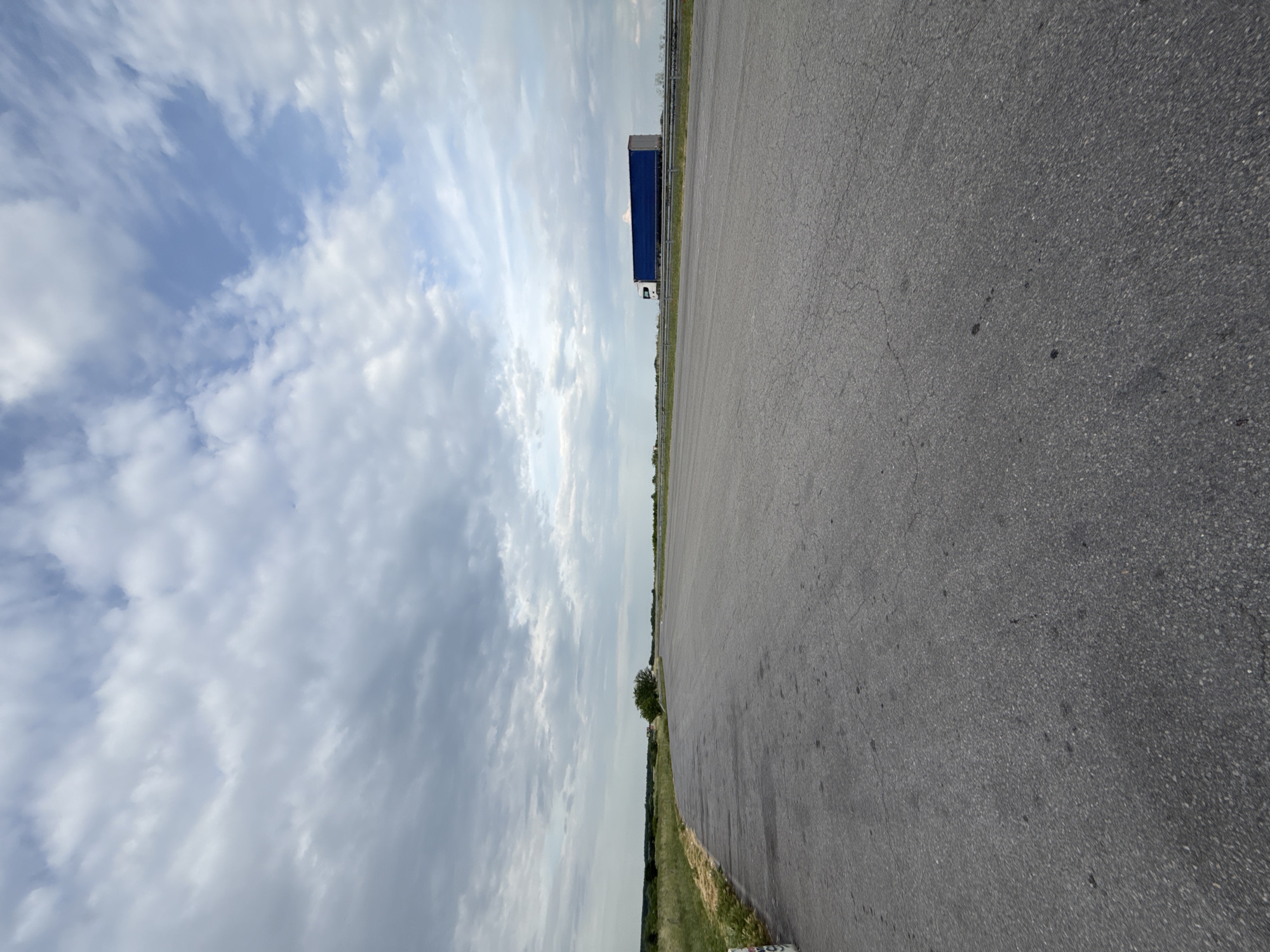 The image shows an asphalt road stretching into the distance under a cloudy sky. A large blue trailer truck is visible on the road in the distance. The roadside features a grassy area with some trees and vegetation. The location is Kolarovo, Bulgaria.