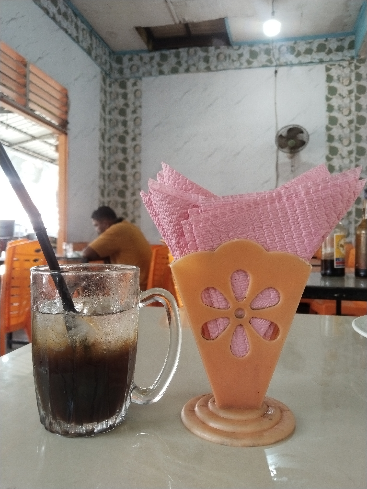 The scene is an interior view of a casual eatery in Meudangara, Indonesia. In the immediate foreground, a clear glass mug containing a dark, iced beverage with condensation on its exterior and a black straw is positioned on a light-colored, reflective tabletop. Adjacent to the mug, to its right, stands an orange plastic napkin holder with a floral cutout design, holding a stack of pink, textured paper napkins.
In the midground, a person wearing a yellow short-sleeved shirt is seated at a dark table, facing away from the camera, with orange plastic chairs surrounding the table. Further right in the midground, another dark table is visible, displaying two bottles, possibly condiments, and a partial white ceramic plate.
The background consists of light-colored walls adorned with decorative green-patterned borders. A single exposed light bulb hangs from the ceiling. A dark-colored wall-mounted ceiling fan is visible on the right wall. The ceiling shows an exposed opening or structural elements. To the left, a large open doorway or portal reveals an exterior area featuring a metal roof structure and ambient daylight.
