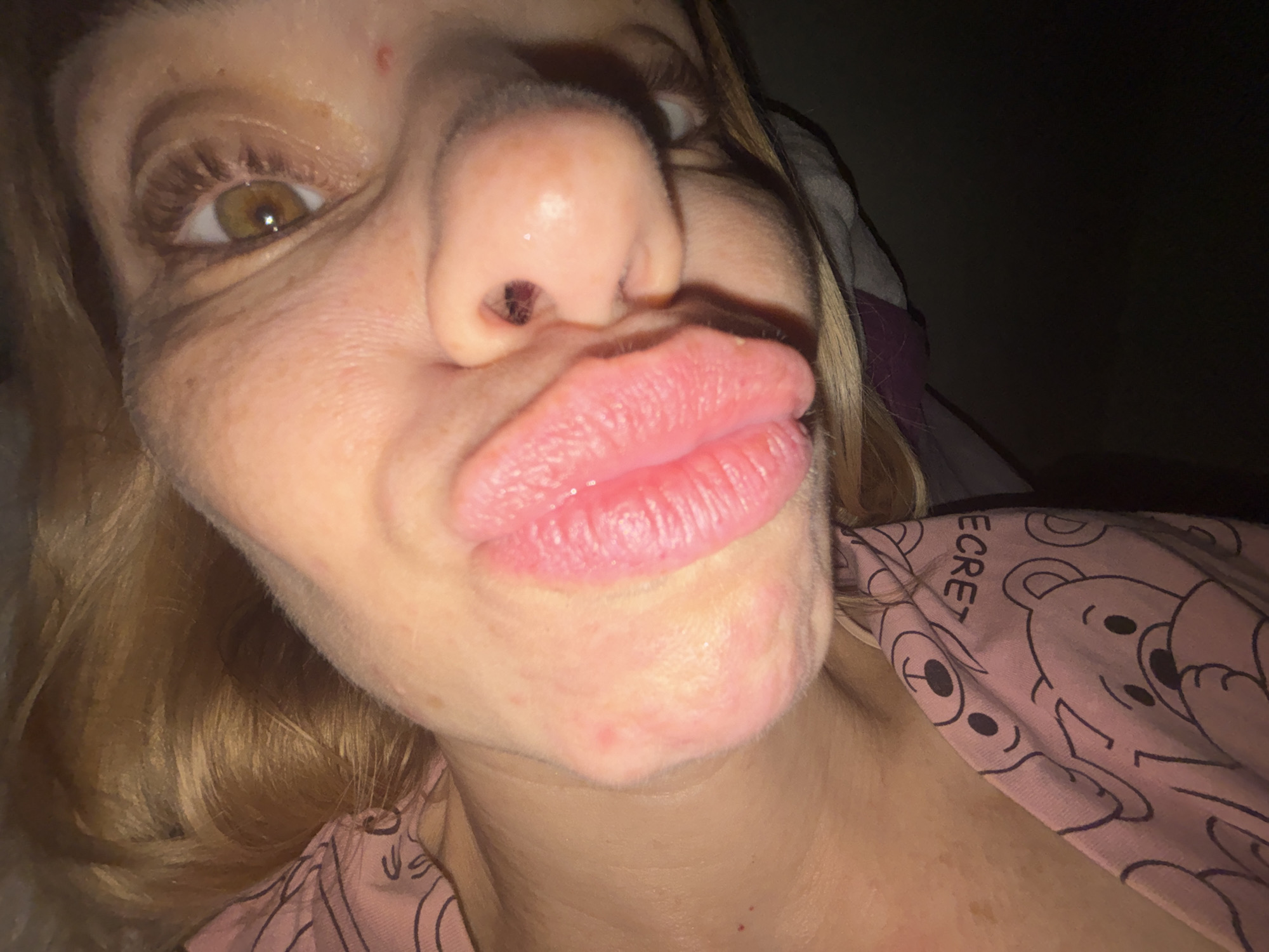 A close-up, selfie-style photograph shows a person's face, with the camera positioned close to their lips, giving a distorted, slightly fish-eye lens effect. The person, appearing to be female with blonde hair, is puckering their lips in a kiss. The lighting is dim, suggesting it might be nighttime or a poorly lit indoor setting.
The focus is primarily on the mouth and nose, with one eye visible on the left side of the frame. The skin appears fair with some freckles. The person is wearing a pink garment with a pattern of cartoon bears and the word "SECRET" visible on it. The background is dark and out of focus, with hints of what might be a pillow or blanket. There are no clear indications of the location, weather, or a specific event taking place. The expression appears playful or flirtatious. The location context provided is Sinyavino, Russia, but this is not visually discernible from the image itself.