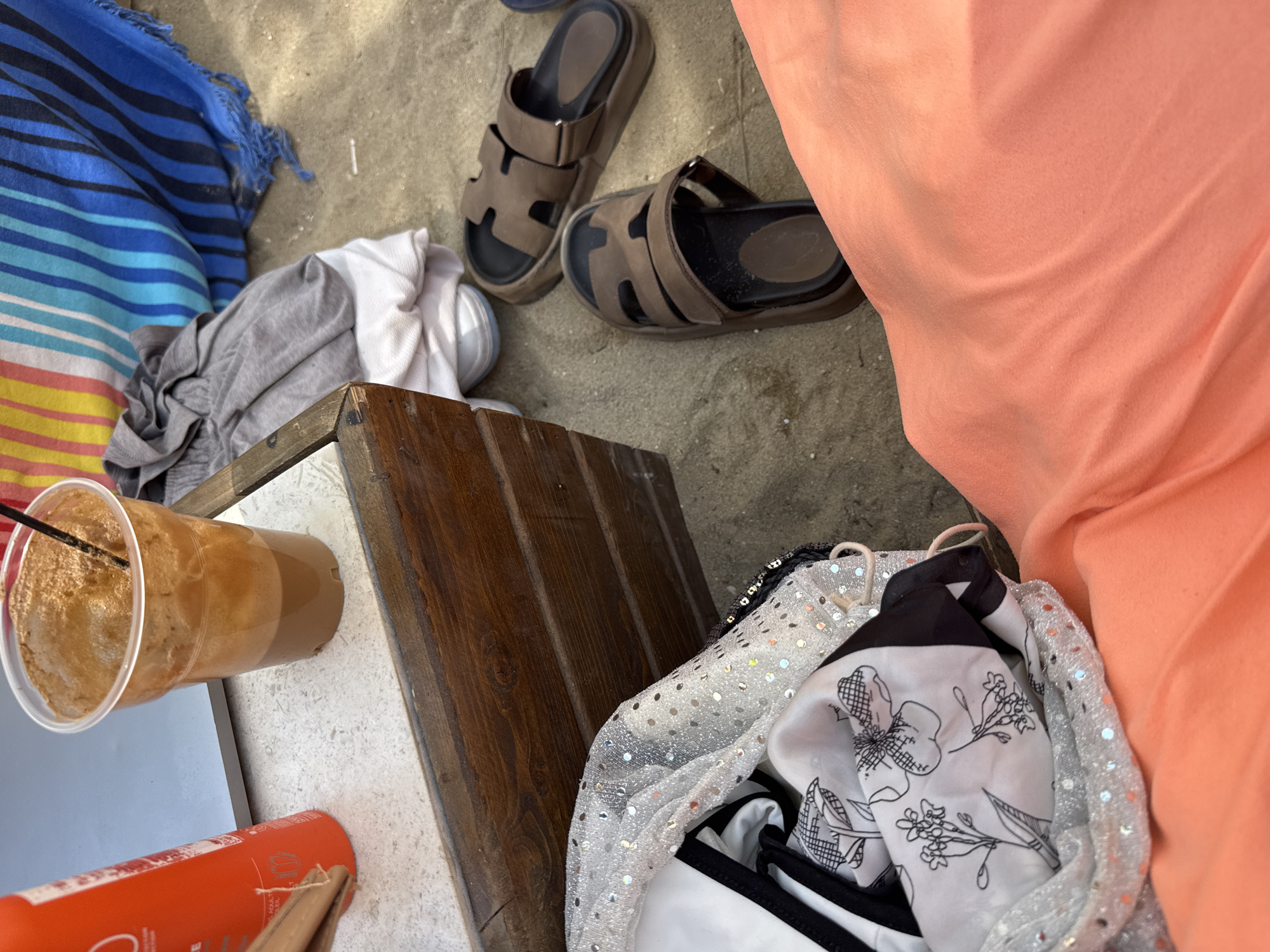 In Thassos, Greece, on a sandy beach, a collection of items are casually arranged. In the foreground, a person's coral-colored clothing is draped over the sand. To the left, a white and black patterned fabric, possibly a scarf or garment, is partly visible, adorned with sparkling silver sequins. Beside it, an orange bottle stands upright. A clear plastic cup filled with a frothy, brown beverage, likely a coffee drink, rests on a textured, light-colored surface, possibly a table or bench, with a dark straw protruding from the top. Next to the cup is a wooden structure. A grey t-shirt and a white cloth are also present. In the background, a striped beach towel in shades of blue, white, yellow, and pink lies spread out. Two dark brown sandals are placed on the sand near the wooden structure and the clothing. The scene evokes a relaxed, seaside atmosphere.