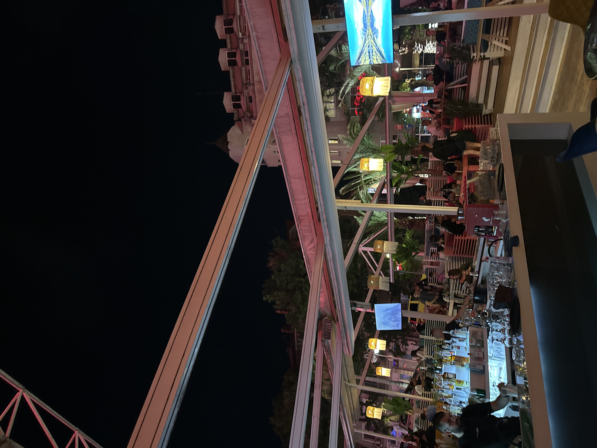 This night-time image captures an outdoor bar or restaurant in Plovdiv, Bulgaria. A white metal framework, likely part of a retractable roof system, spans the upper portion of the scene, reflecting subtle red and pink ambient lighting against the dark, clear sky.
In the foreground, a white, illuminated bar counter is visible, behind which a male bartender with a beard, wearing a dark t-shirt, is actively preparing a drink, holding a shaker. Numerous bottles of various spirits are arranged on backlit shelves behind him, alongside stacks of glassware.
Beyond the bar, the venue features tiered seating areas with white benches and tables. Multiple groups of people, including men and women, are seated and standing, engaging in conversations and socializing. One individual is leaning towards the bar, seemingly interacting with staff.
Decorative elements include several hanging lamps with woven, cylindrical shades emitting warm yellow light, and potted palm trees and other green foliage. Two flat-screen monitors are mounted within the overhead structure; the screen on the right displays a brightly colored, symmetrical image of a mountain reflection on water, while the screen on the left shows a less distinct visual.
In the background, multi-story buildings of Plovdiv are illuminated, including one with a distinctive pointed roof and multiple dormer-like structures. Glimpses of a street with distant lights and a yellow vehicle are visible further back.