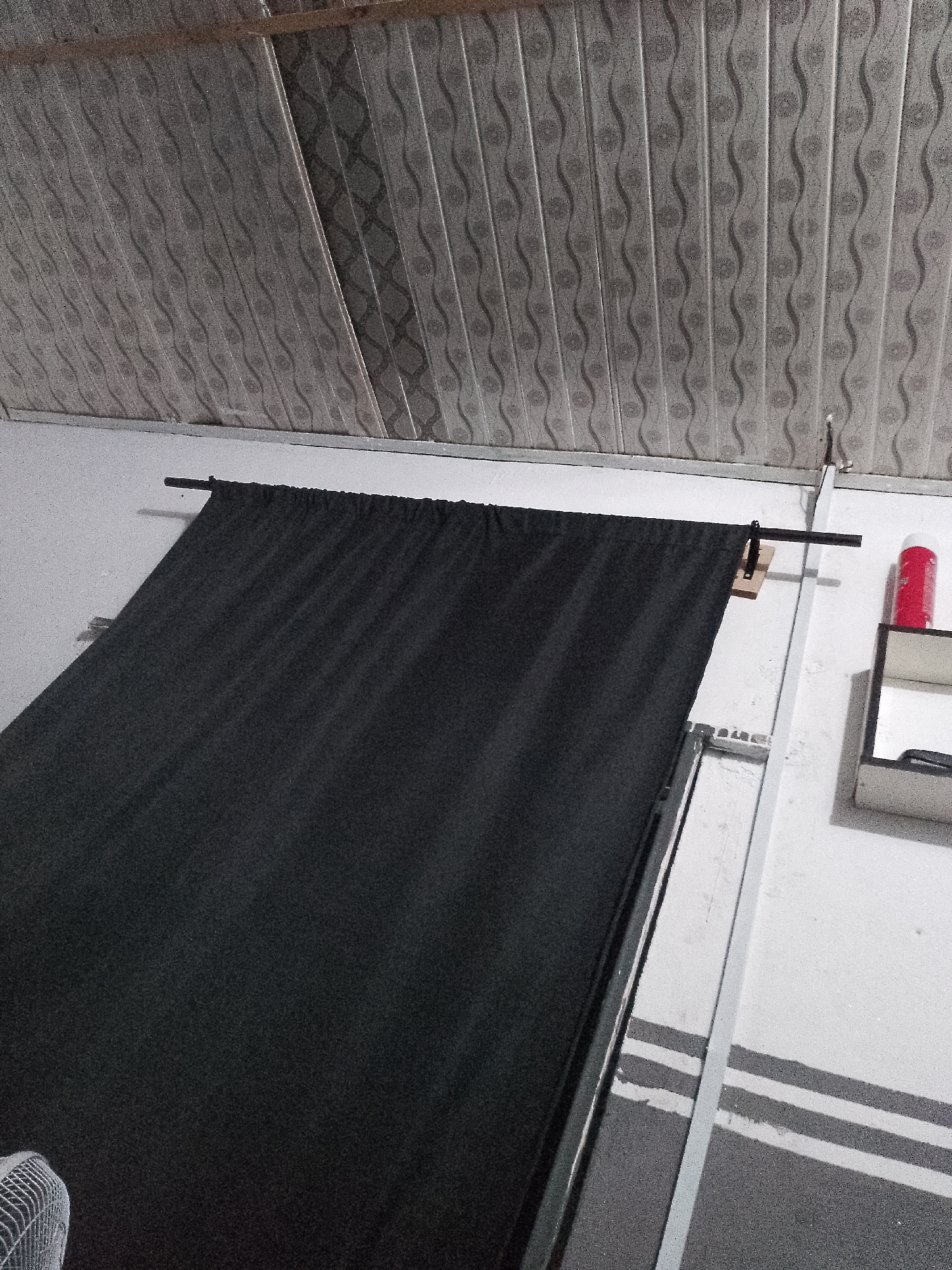 The image shows a dark fabric hanging from a rod attached to a wall. The ceiling above is covered with textured panels that have a repeating wavy pattern. A white vertical support beam is visible on the right side of the image, and a small shelf with a red object and a dark object is mounted on the wall next to it. The lower part of the image shows a dark surface with white stripes and a portion of a fan on the left. The scene appears to be indoors, possibly a room or a studio. The location is Unknown, Unknown.
