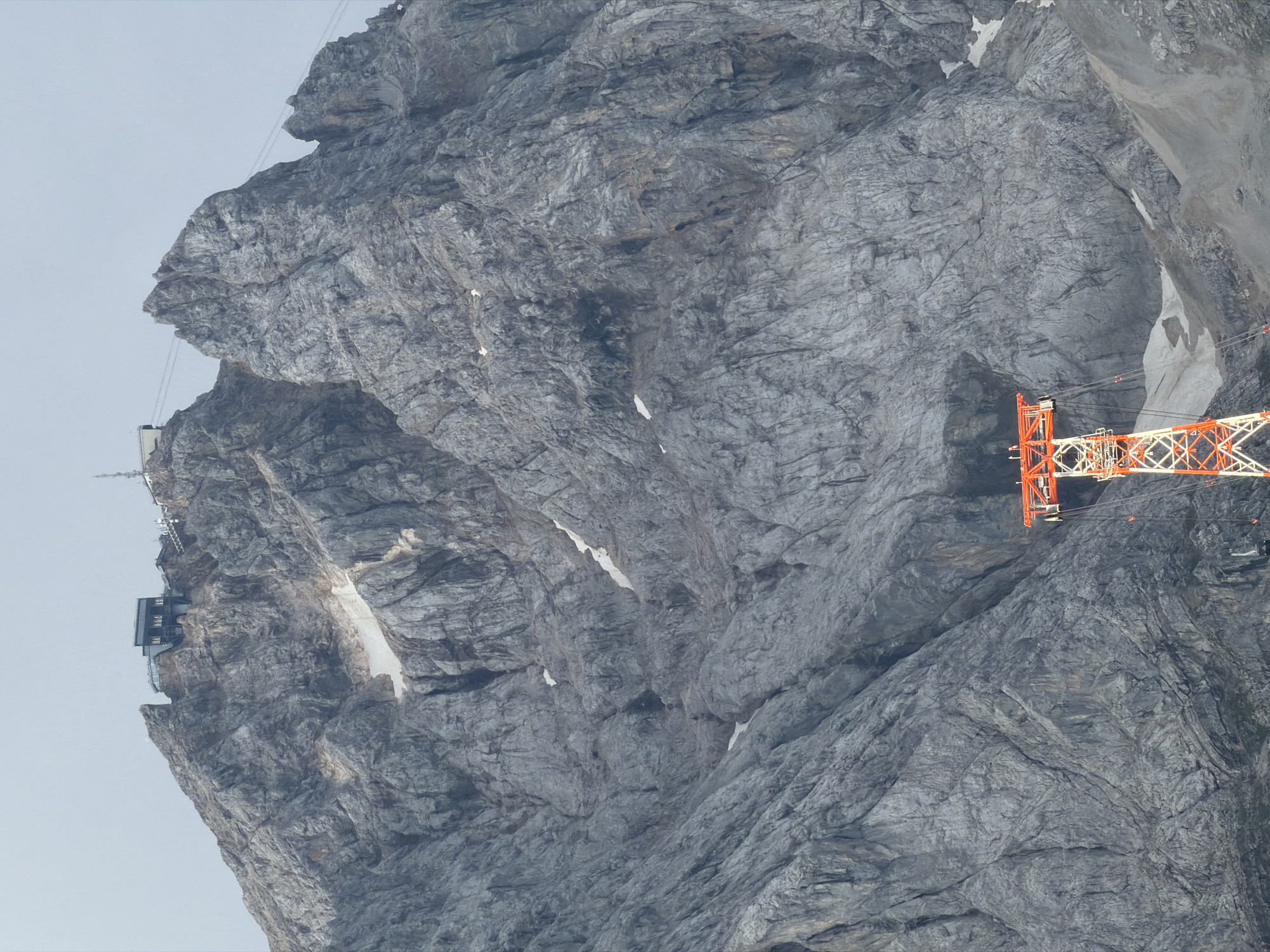 This is a photograph of the Zugspitze, Germany's highest peak, located near the municipality of Grainau. The image captures a vast, craggy mountain face, predominantly gray and textured with numerous rock formations and crevices. Patches of snow are visible scattered across the slopes, indicating a high altitude and a potentially cold environment.
Dominating the lower right portion of the frame is a bright orange and white pylon, part of a cable car system. The pylon is a tall, lattice-like structure with intricate metalwork. Cables extend from the top of the pylon, disappearing into the mountainous terrain, suggesting it is a support for a gondola or cable car line.
Further up the mountain, near the summit, are man-made structures. These include buildings with dark roofs and large windows, as well as what appear to be communication towers or antennas. The presence of these structures indicates a developed area on the peak, likely for tourism or scientific observation.
The sky is overcast with a light gray hue, suggesting a cloudy day. The overall impression is one of a dramatic, rugged natural landscape with technological infrastructure integrated into its summit. No people are visible in the image.