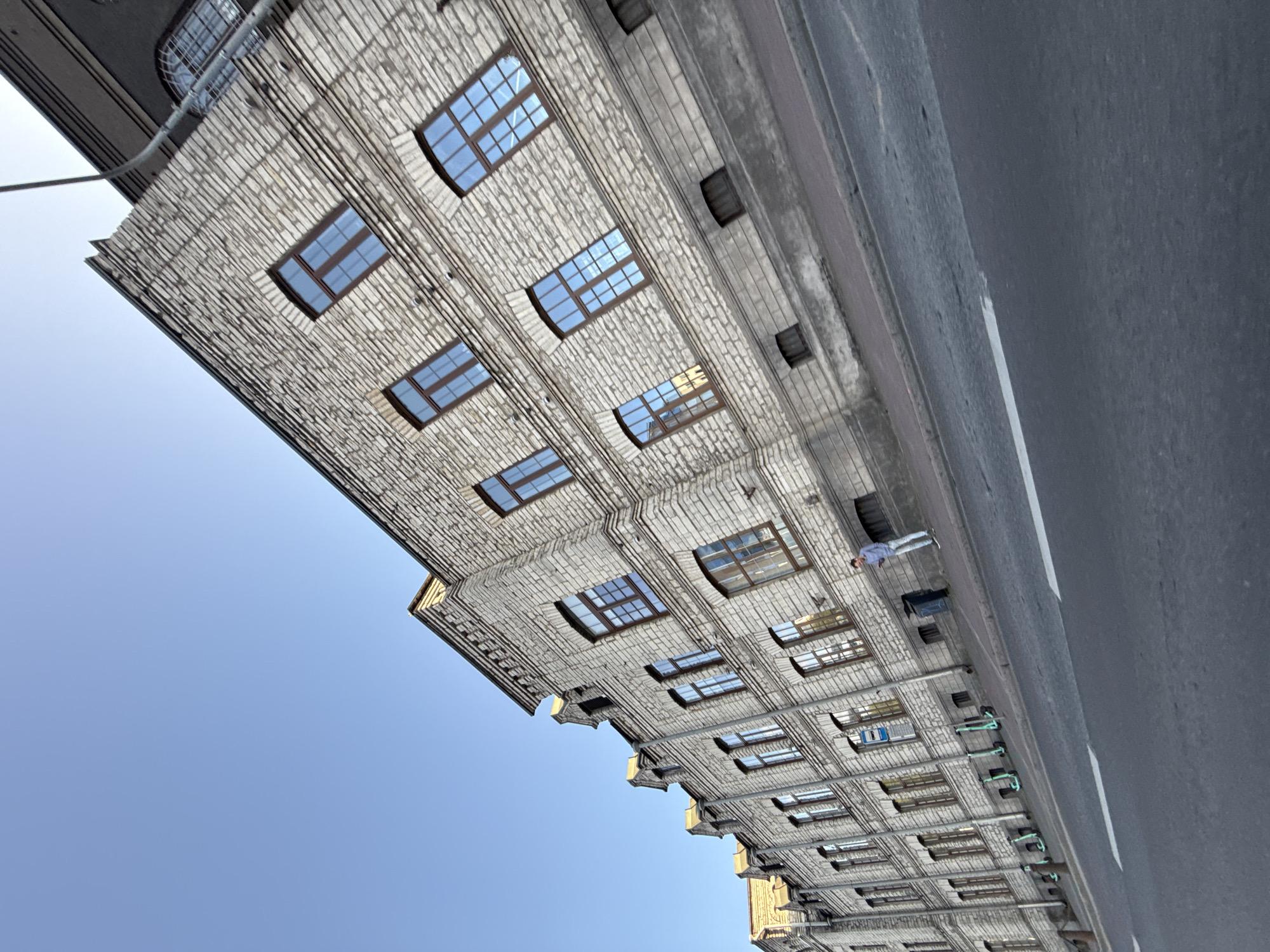 The image shows a long, multi-story building with a light-colored brick facade and numerous windows with dark frames. The building appears to be located on a street in Tallinn, Estonia, as indicated by the distinctive architecture and the presence of several electric scooters parked along the sidewalk. The sky is clear and blue. A single person, a male wearing a light blue shirt and dark pants, is standing on the sidewalk looking at a phone. The street in the foreground has a white line marking a lane. The perspective of the image is tilted, giving a dynamic view of the scene.
