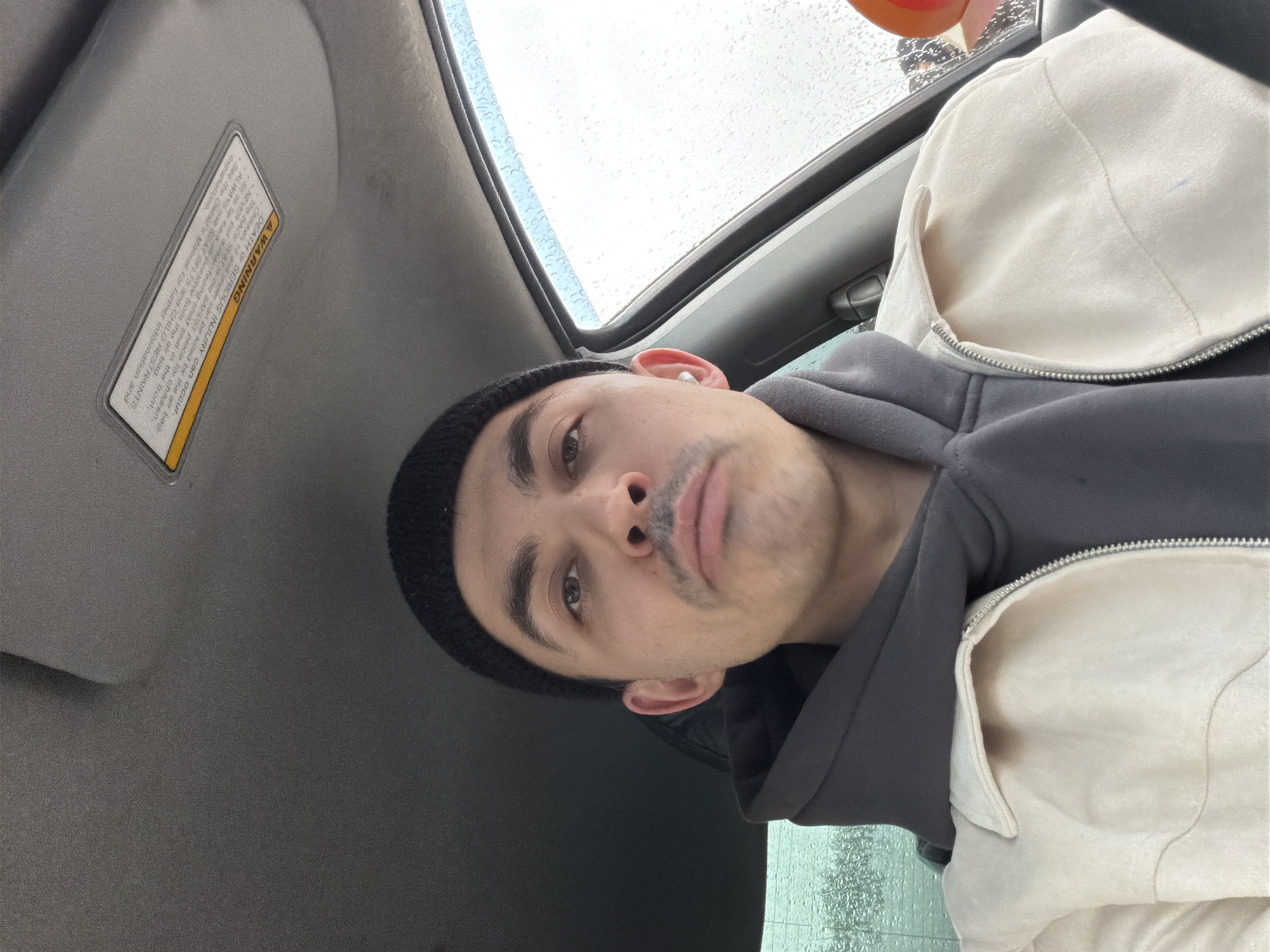 A close-up, slightly angled, selfie of a young adult man, positioned inside a vehicle, possibly a car, looking directly at the camera. He is wearing a dark-colored beanie, a grey hooded sweatshirt, and a light-colored jacket. His face is clean-shaven except for a thin mustache. Part of the vehicle's interior is visible, including the sun visor and a portion of what appears to be the sunroof. Raindrops are visible on the sunroof glass. A warning label is affixed to the sun visor. The background outside the vehicle is blurred due to the raindrops on the glass. The location is not discernable from this image, so stating that it is in Izhevsk, Russia, is impossible.
