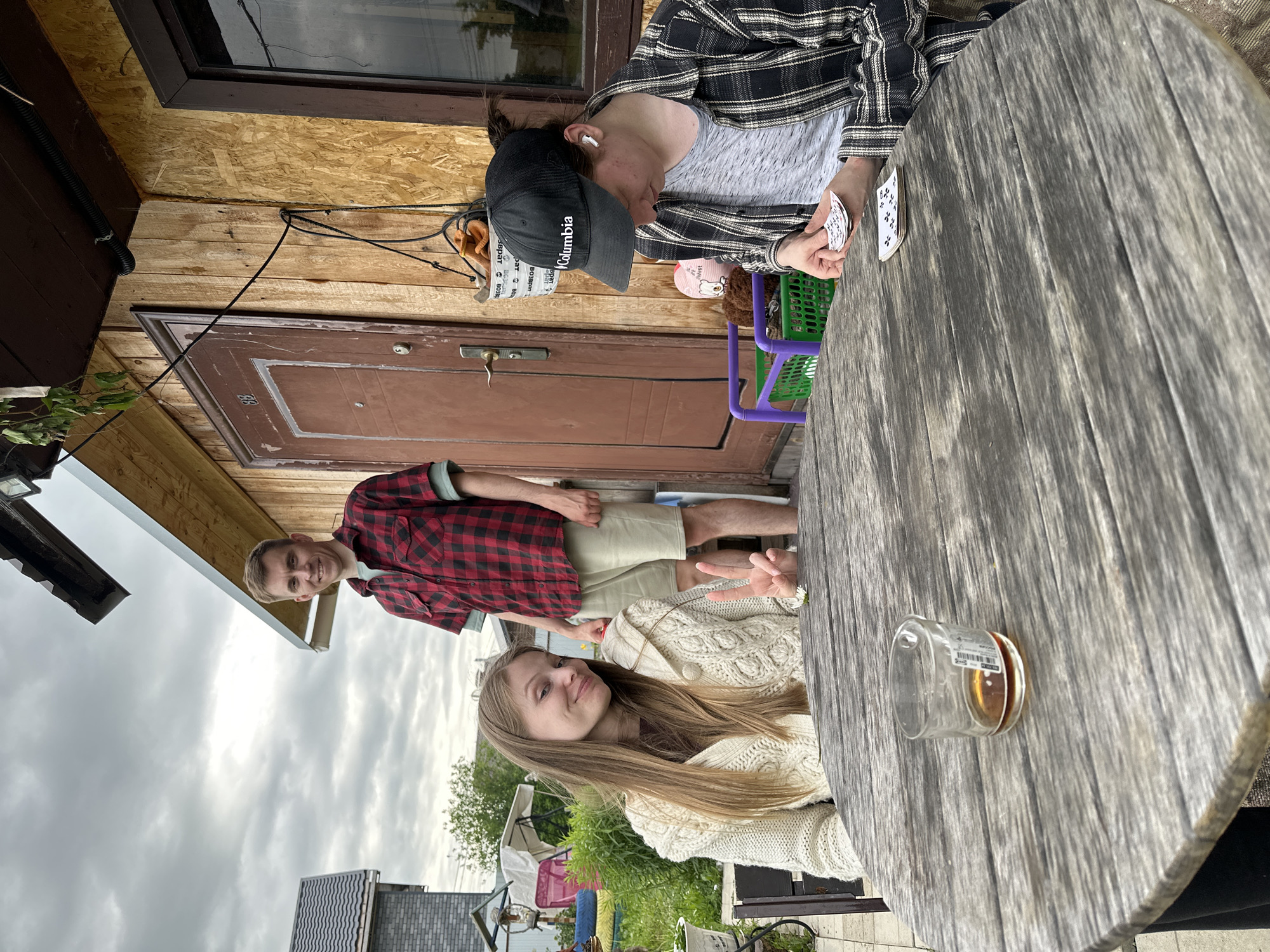 This image captures an outdoor scene featuring three young adults in a backyard setting in Bol'shaya Mos', Russia, under an overcast sky. In the foreground, a large, weathered round wooden table occupies the center of the frame. On the table, to the left, is a clear glass tumbler containing a small amount of amber liquid, with a white sticker adhered to its side. A small white daisy-like flower rests on the table's surface towards the center. Seated at the table on the left is a young woman with long, light brown hair, wearing a cream-colored chunky knit cardigan over a dark top. She is looking towards the camera with a slight smile and forming a peace sign with her right hand. To the right of the table, a person is seated, largely obscured from the chest down. This individual wears a black "Columbia" baseball cap, a grey mottled t-shirt, and a black and white plaid long-sleeved overshirt. White wireless earbuds are visible in both ears. Their gaze is directed downwards, focused on a small stack of white cards with black markings held in their hands, with another small stack resting on the table in front of them. Standing behind the table, slightly to the left of a wooden door, is a young man with short, light brown hair, wearing a red and black plaid short-sleeved shirt over a light green t-shirt, and light-colored shorts. He is smiling and looking towards the camera. The background features a rustic building, likely a private residence or dacha. The exterior is composed of vertical wooden planks on the left side of the door and Oriented Strand Board (OSB) sheets to the right. A dark brown door with the number "88" is prominent, featuring a modern handle and lock. To the right of the door is a dark-framed window. Above the door and window, a dark brown wooden soffit extends, under which a black corrugated conduit pipe is visible. A black cable runs diagonally across the wooden facade from the upper left. A white bucket-like container, with partially visible text that includes "ода", hangs from the wall near the door handle and contains brown, soft objects. Below the window, a purple plastic crate and a green plastic basket are present, alongside a brown plush toy. Further in the background, to the far left, a grey brick house with a dark tiled roof is partially visible.
