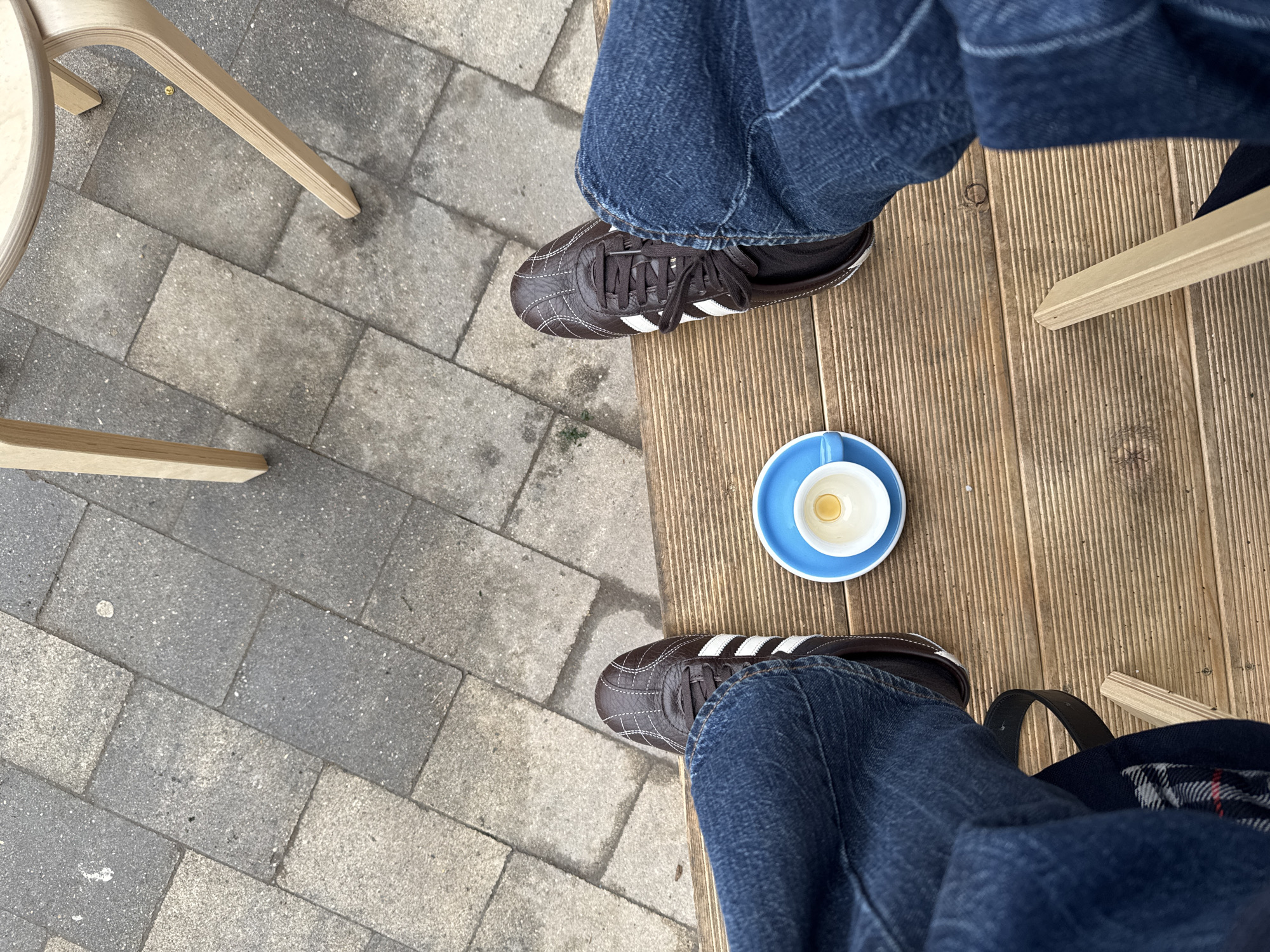 The photograph captures a scene at an outdoor café. Two chairs are visible, one positioned on a gray-paved area, the other on a wooden platform. A person wearing blue jeans and brown leather shoes with white stripes is seated. Between the person's feet, on the wooden platform, sits a light-blue saucer with an empty white coffee cup. The person's position and the presence of the café setting suggest a casual, relaxed moment. The location of Moscow, Russia, cannot be confirmed with the given information.