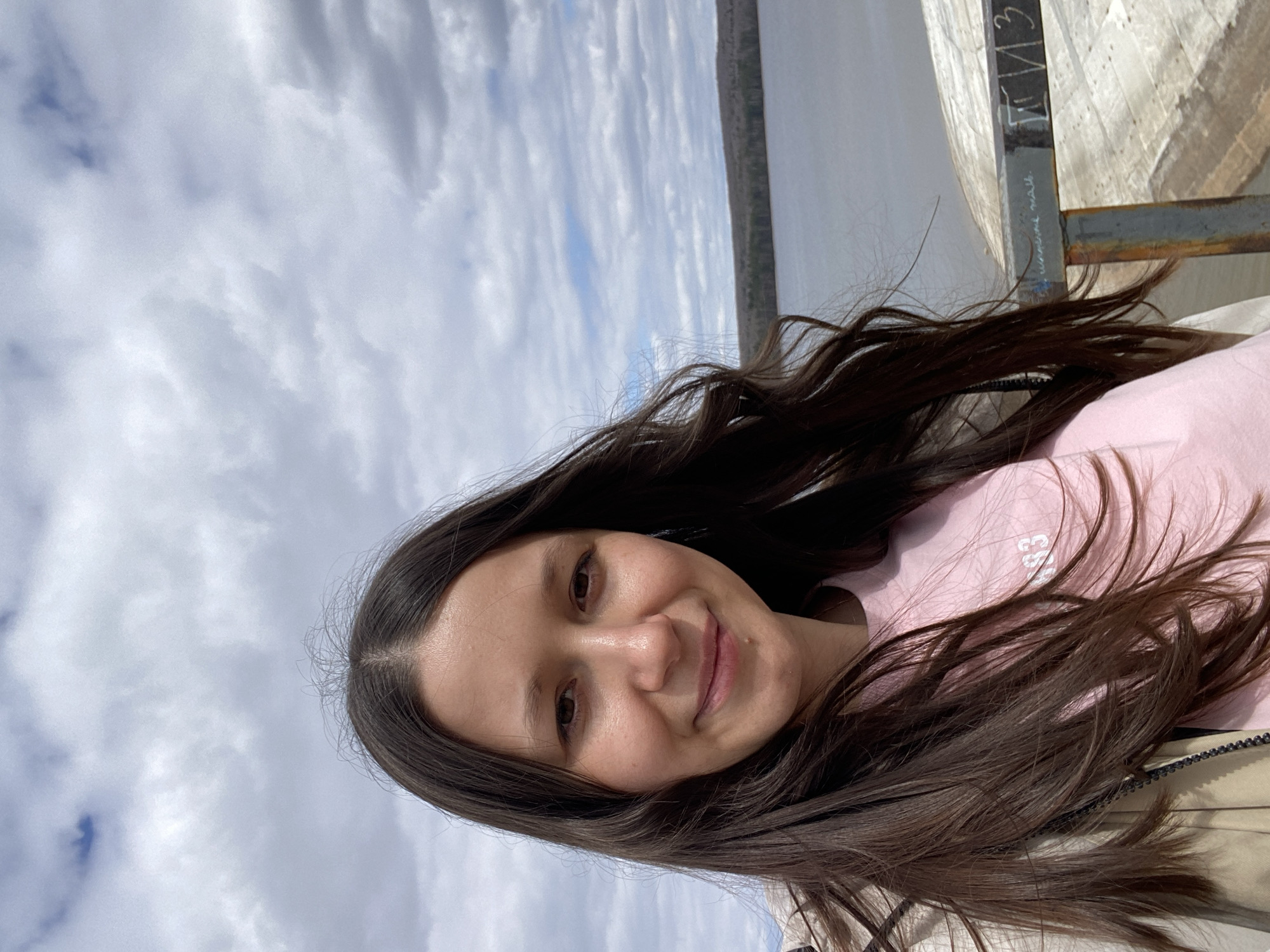 A selfie of a young woman with long, dark brown hair, wearing a light pink sweatshirt and a beige jacket, taken outside on a cloudy day. The background includes a body of water and a grassy shoreline. A metal structure is visible to the right. The location appears to be in Chaikovsky, Russia, based on the inscription on the metal structure which appears to be written in Russian.