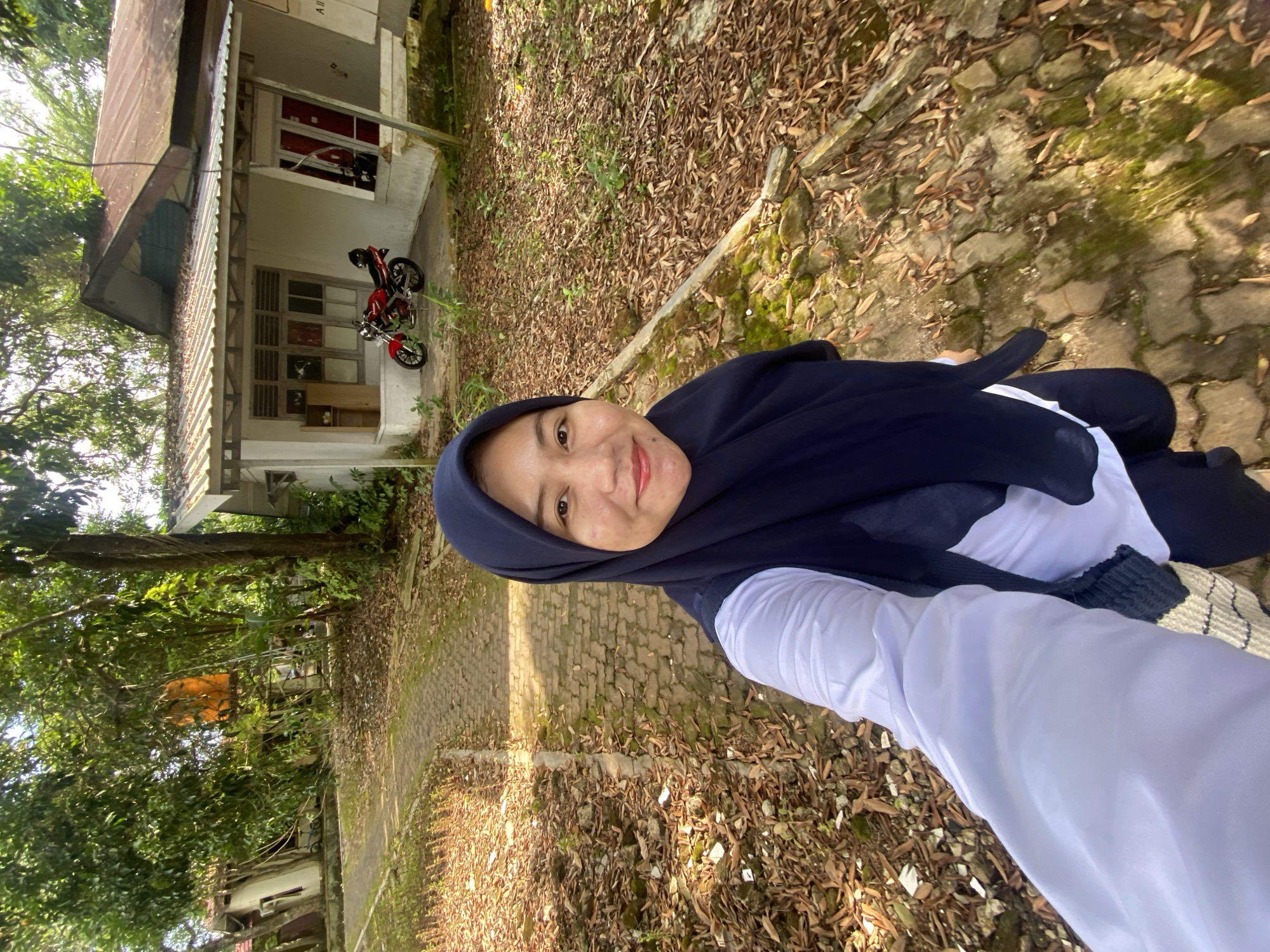A woman wearing a dark blue hijab and a white shirt stands on a paved path lined with dry leaves and moss-covered stones. She is taking a selfie, with her right arm and hand visible in the foreground. Behind her, a red motorcycle is parked in front of a light-colored house with a weathered, reddish-brown roof. The house has several windows and a covered porch area. Tall trees with green foliage surround the area, dappling the ground with sunlight. The scene appears to be an outdoor residential setting in Banjarbaru, Indonesia.
