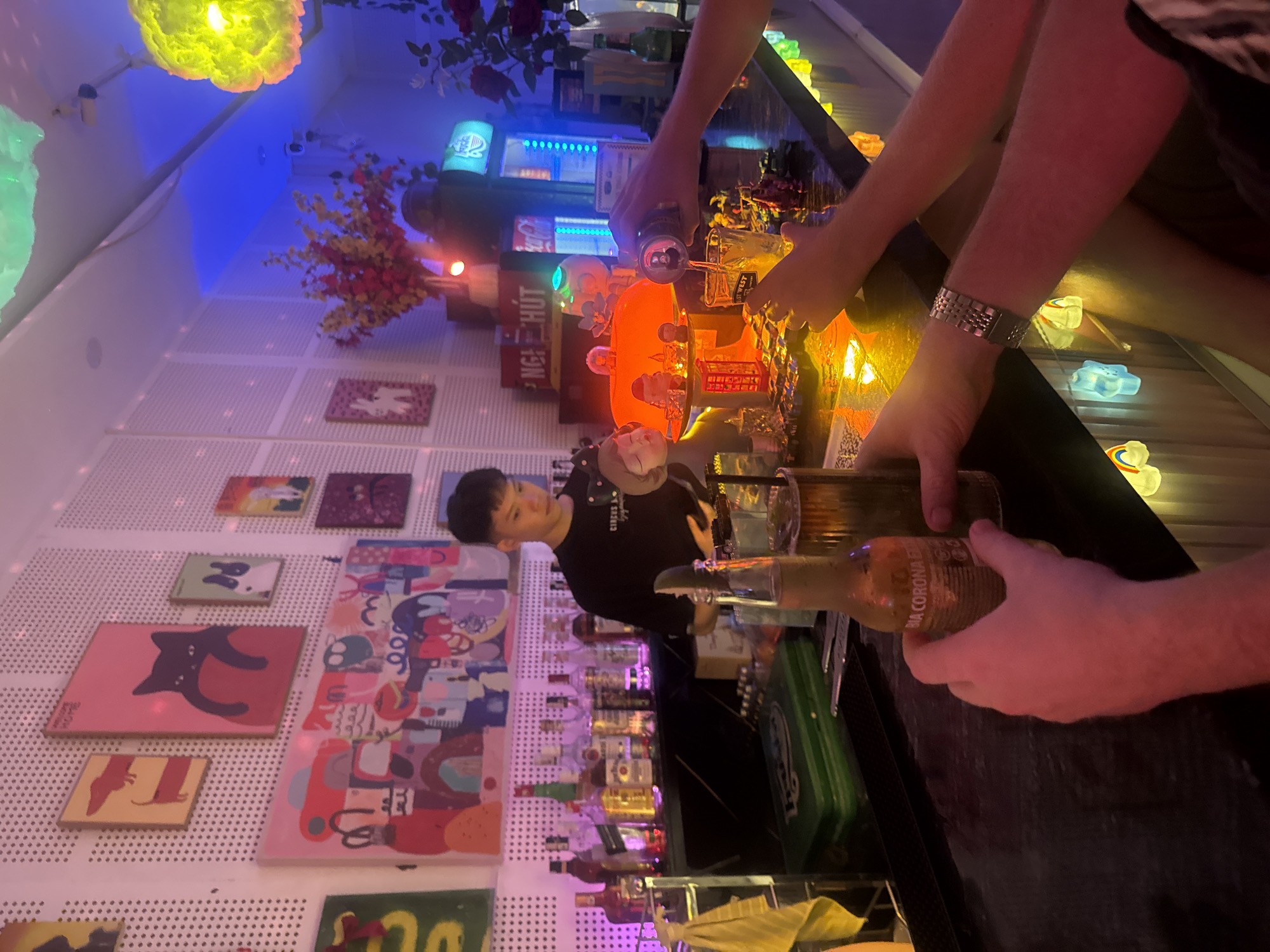 This indoor image, captured from a slightly elevated perspective, is rotated ninety degrees clockwise, depicting a lively bar scene in Đà Nẵng, Vietnam, likely during the evening or night. The setting is characterized by dim ambient light and an abundance of colorful decorative lighting and whimsical decor.
Three individuals are visible: a male bartender with dark hair, wearing a black t-shirt (partially reading "CIRCUS A"), stands behind the bar, looking towards the right. In the foreground, two sets of hands are present. One pair, adorned with a metallic watch on the left wrist, holds a can and pours liquid into a glass (partially visible text: "WEST"). The other pair holds a bottle labeled "BIA CORONA ES" (Bia Corona Extra).
The bar counter is dark, cluttered with bottles, glasses, and decorative elements such as small, glowing star and cloud-shaped lanterns, a miniature red phone booth, and a white skull with glowing green eyes. Behind the bartender, shelves are stocked with various bottles of spirits. The background wall is light-colored and textured, adorned with numerous framed artworks featuring abstract designs, colorful patterns, and figures, including a prominent black cat illustration. Artificial red and yellow flowers add another decorative touch.
The atmosphere is vibrant and festive, enhanced by the neon lights, including a sign with partially visible Vietnamese text "NGA HUT," which serves as a location cue. The activity centers around drink preparation and serving, with both bartender and patrons engaging with beverages.