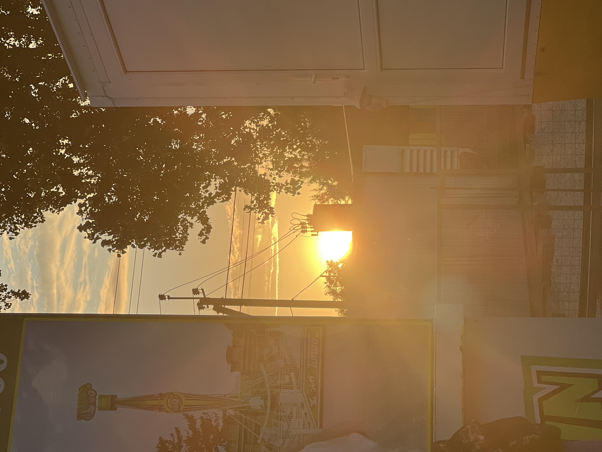 The image depicts a sunset scene in Golubitskaya, Russia. In the foreground, a large advertisement panel features a graphic of a tall amusement park ride. To the right, the side of a white structure, possibly a trailer or kiosk, is visible. In the background, the sun is setting, casting a warm golden glow across the sky. Silhouetted against the bright light are trees and utility poles with power lines. The overall impression is one of a relaxed, late afternoon atmosphere at what appears to be an amusement or fairground.