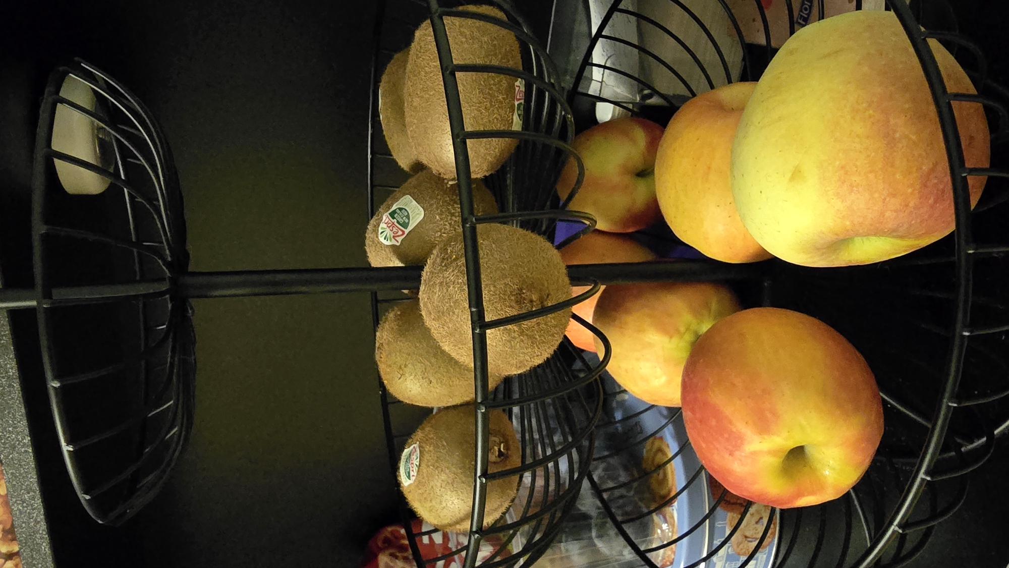 The image shows a black wire tiered fruit basket containing several fruits. The top tier holds several kiwis, some with "Zespri" stickers. The middle tier also contains kiwis. The bottom tier contains several apples, with red and yellow coloring. The fruit basket is placed on a dark, textured surface, likely a countertop or shelf. The background is dark and out of focus, suggesting an indoor setting. There are no people or animals visible in the image. The lighting is dim, highlighting the fruits in the basket. The location of this image is unknown, and there is no information about the city of Neuwied, Germany.