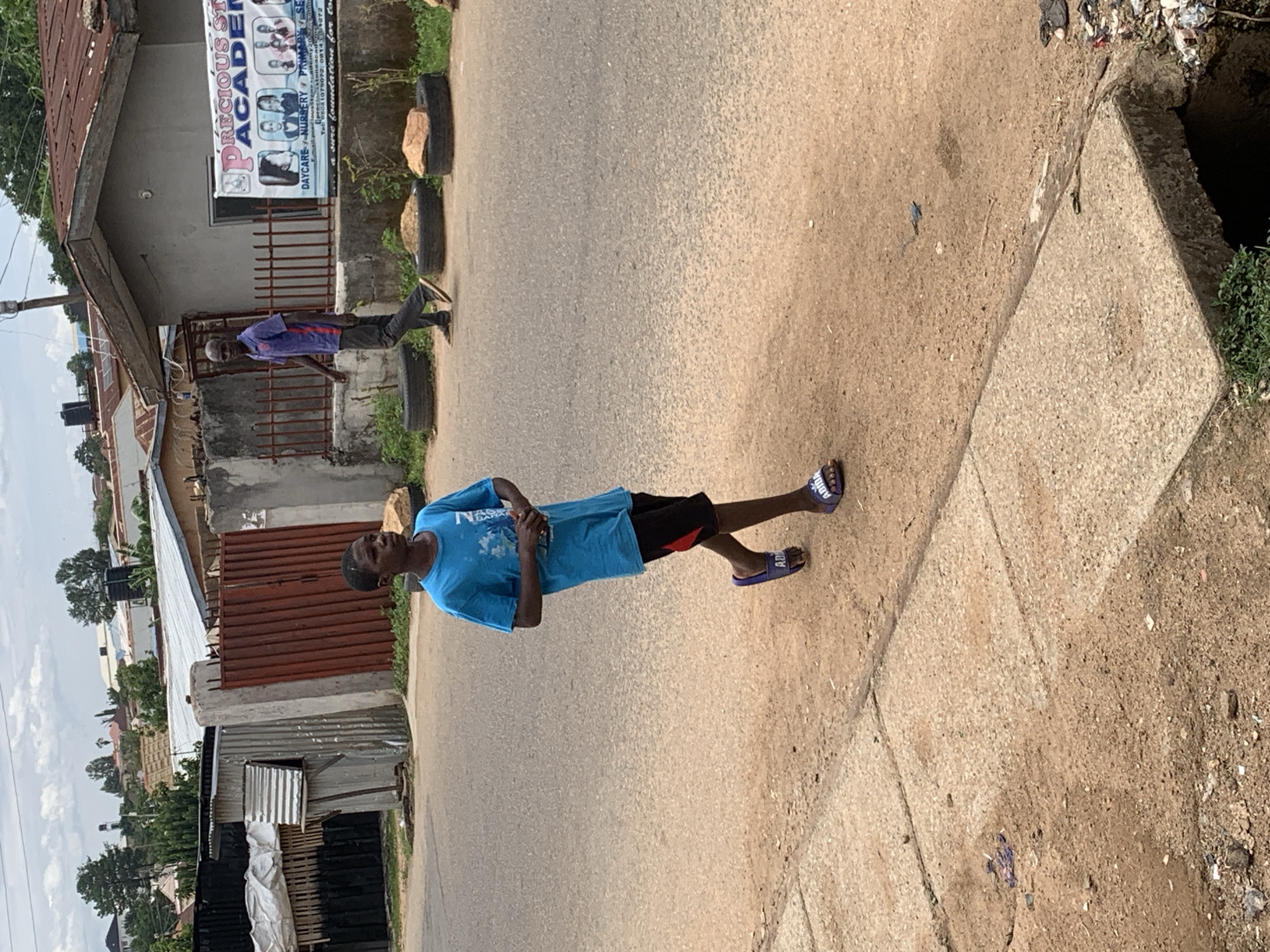This image captures an urban street scene in Jos, Nigeria, under an overcast sky. The foreground features a young dark-skinned male, likely a teenager, walking leftward on a dirt-covered asphalt road. He wears a bright blue short-sleeved t-shirt, black shorts with a red side stripe, and purple Adidas slide sandals, with his hands clasped in front. In the mid-ground on the right, an adult dark-skinned male also walks leftward, attired in a purple polo shirt and dark trousers, with light-colored sandals.
The background on the left reveals several structures constructed from corrugated metal sheets, some appearing to be shanties or ancillary buildings, and a prominent rust-colored metal gate forming part of a compound entrance. Beyond these, additional buildings with metallic roofs are visible on a slight incline amidst sparse foliage. On the right, a single-story building with a reddish-brown corrugated roof and a light-colored facade is present. A large white banner for "Precious Stone Academy" is affixed to its fence, advertising daycare, nursery, and primary school services. Three black car tires, two containing soil and one with a reddish rock, line the edge of the road in front of the academy. The road surface consists of a mix of asphalt and packed earth, bordered by a concrete sidewalk on the left and a concrete-covered drainage ditch containing visible refuse on the far bottom right.
