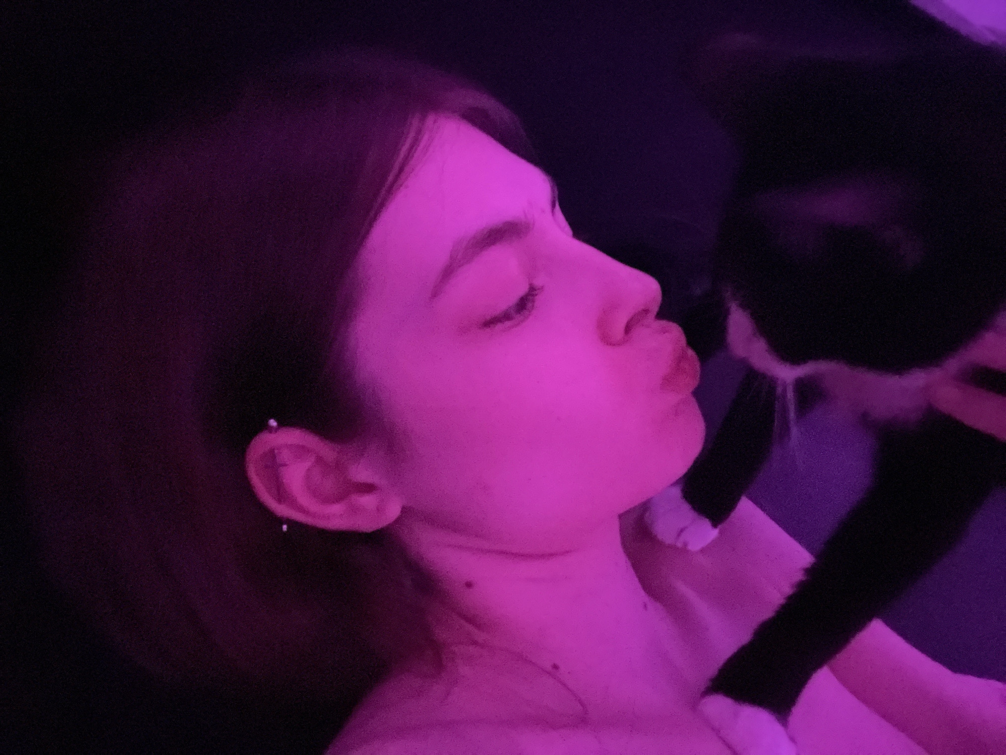 An intimate portrait features a young woman in profile on the left, illuminated by strong pink and purple ambient lighting, as she interacts affectionately with a black and white cat on the right. The woman, with dark hair, has her lips pursed into a kiss, directed towards the cat. Her right ear is visible, adorned with multiple piercings.
The cat, a tuxedo breed with black fur and white markings on its chest and paws, is positioned close to the woman's face. One of its white paws rests gently on the woman's shoulder or upper chest. The interaction suggests a moment of tenderness and connection between the two.
The setting appears to be an indoor space, likely a dimly lit room, with the dark background subtly catching the purple light, creating a cozy and warm atmosphere, indicative of evening or nighttime. The overall lighting casts a distinct magenta hue over both subjects. There is no visible text in the image. The scene takes place in Saint Petersburg, Russia.
