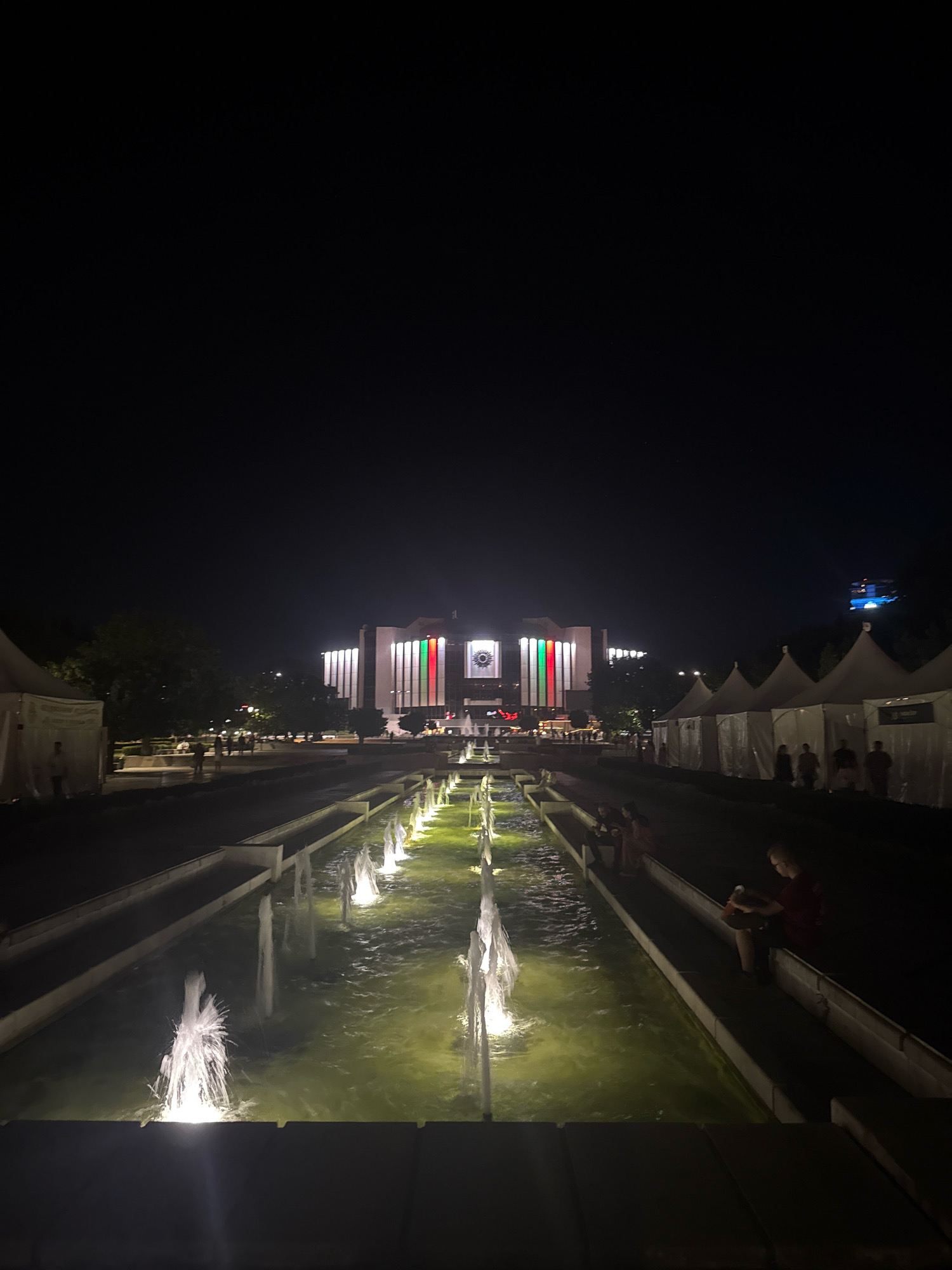 At night, the National Palace of Culture in Sofia, Bulgaria, is illuminated, showcasing its prominent facade. The building is adorned with vertical bands of green, white, and red lights, representing the Bulgarian flag. In front of the palace, a long, shallow fountain stretches across the scene, with multiple water jets illuminated from below, casting a greenish glow on the water. Several white tents are set up on the right side of the image, possibly for an event or exhibition. People are visible in the foreground, some seated along the edge of the fountain and others walking in the distance near the tents and the palace. The sky is dark and clear, indicating nighttime.