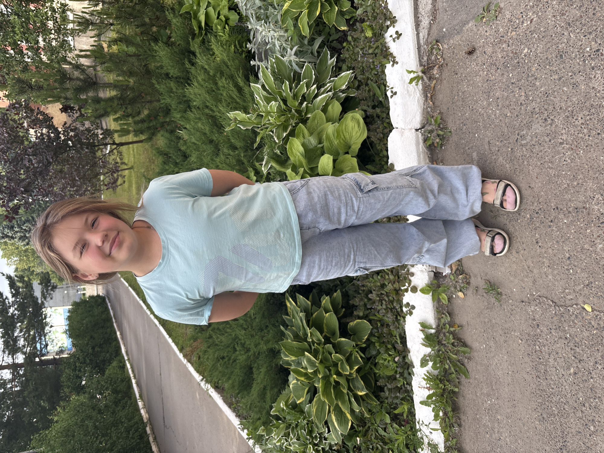 A young girl with blonde hair, wearing a light blue t-shirt and light gray cargo pants, stands in front of a landscaped area in Chaikovsky, Russia. She is looking at the camera with a slight smile. The girl is positioned on a paved walkway next to a garden bed filled with lush green hostas and other plants. A white concrete border separates the walkway from the garden. In the background, there are trees, bushes, and a building. The overall scene appears to be an outdoor public space, possibly a park or a residential courtyard.