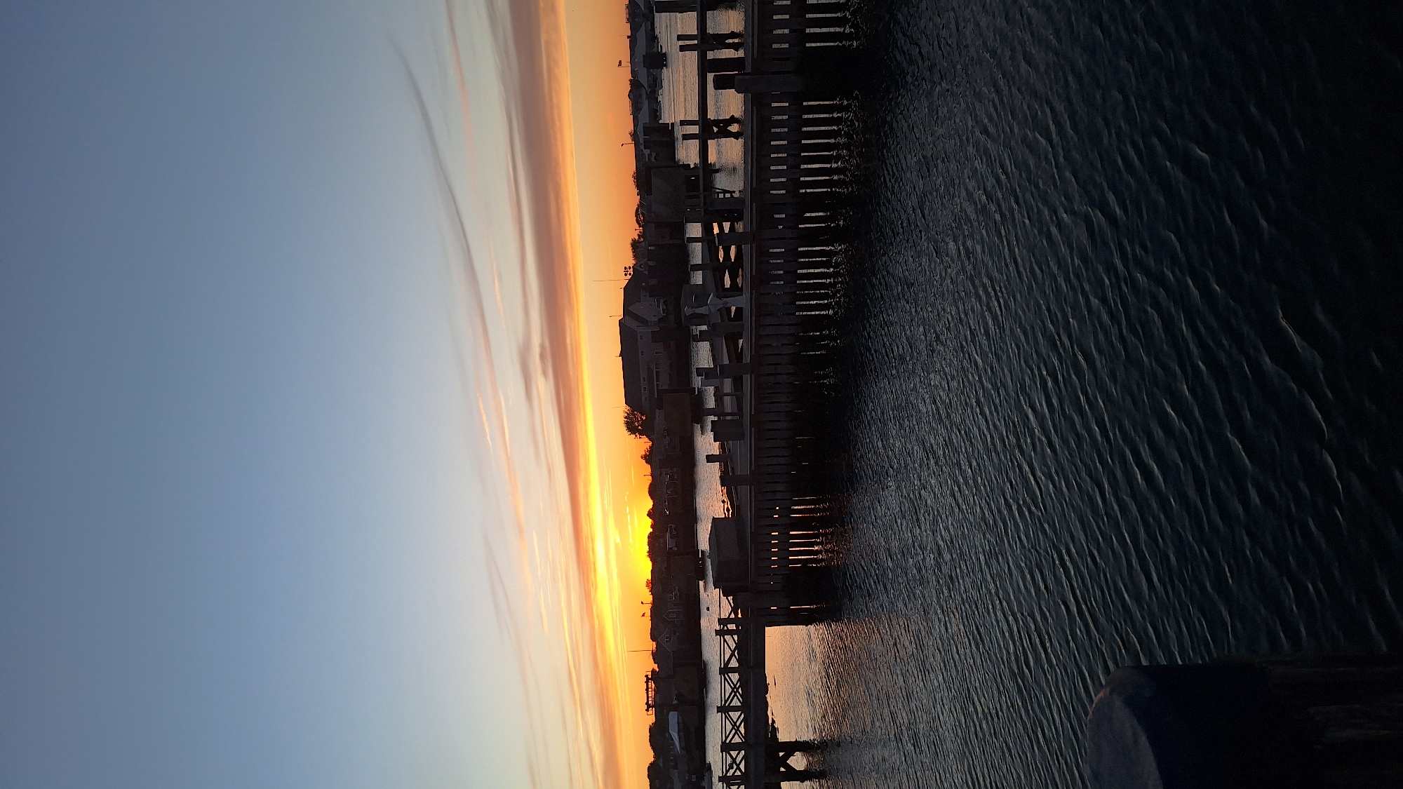 This is a photograph of a harbor at sunset. The sky is a gradient of soft blues and vibrant oranges, with wispy clouds stretching across the horizon. The water is dark and rippled, reflecting the warm hues of the sunset.
In the foreground, a dark wooden pier juts out into the water, with several boats docked alongside it. The pier is weathered and worn, suggesting it has been here for a long time. The boats are silhouetted against the colorful sky, their masts and rigging creating a delicate pattern against the sunset.
In the background, a line of houses and buildings stretches along the shoreline, their dark shapes contrasting with the bright sky. A few trees are scattered among the buildings, their branches bare against the fading light. The overall impression is one of peace and tranquility, a quiet moment as the day comes to a close. The location is likely a coastal town or village in Nantucket, USA, given the presence of a harbor and the distinct architectural style of the buildings. The time of day is clearly sunset, indicated by the colors of the sky and the low angle of the light. The weather appears to be calm, with no indication of strong winds or storms.