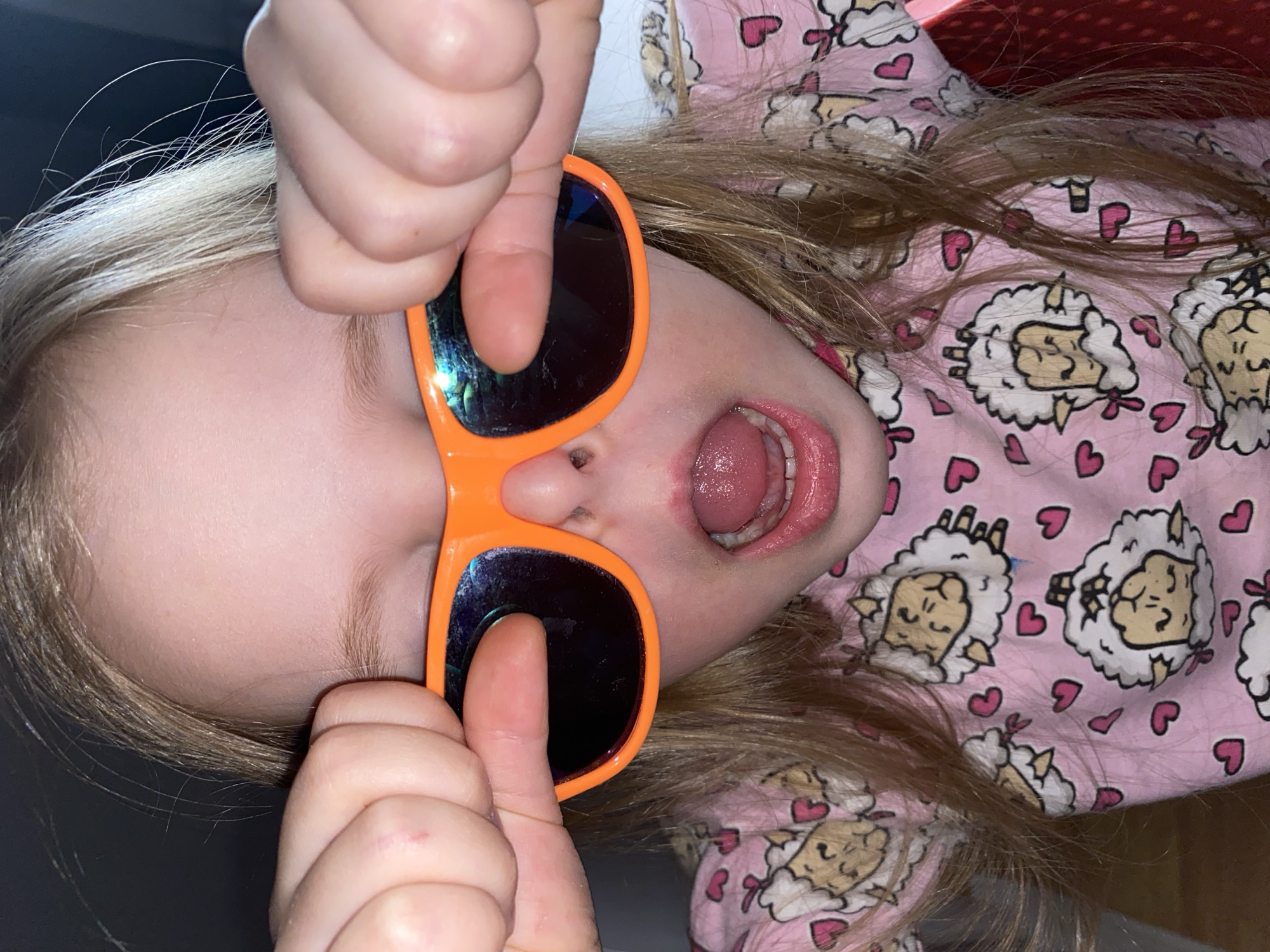 A young child with blond hair is lying on their back, wearing orange sunglasses and a pink pajama top printed with sheep and hearts. The child's mouth is open, and they are holding the sunglasses to their face with their fingers. The location is not discernible from the image, and there is no indication it is in Votkinsk, Russia.