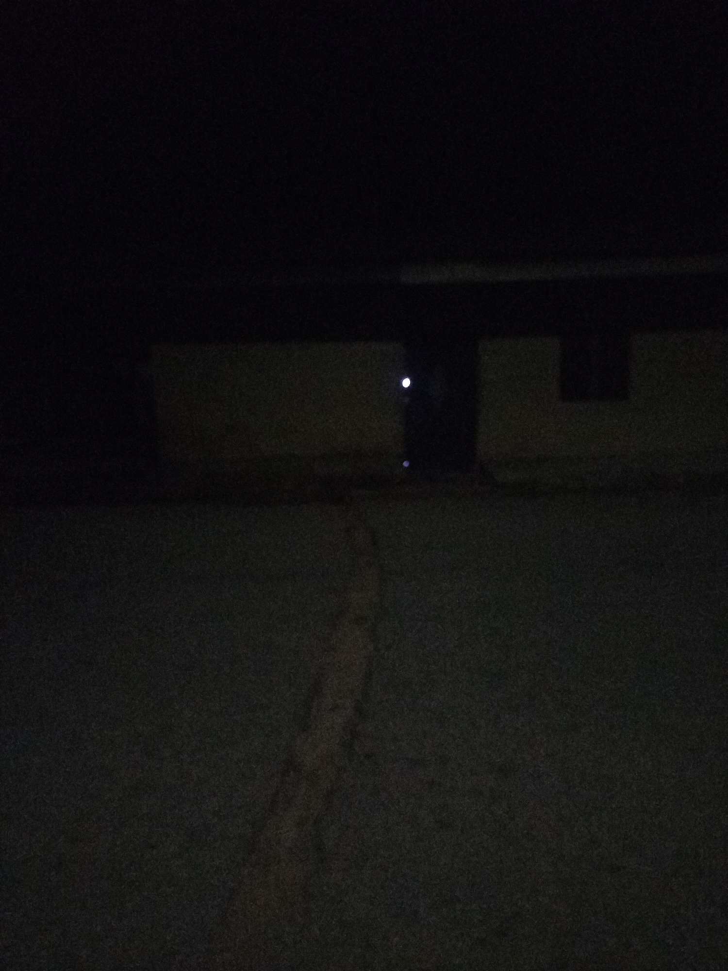 The image depicts a dark, grainy scene at night, likely in the city of Jalingo, Nigeria. In the background, a low-rise building is visible with dimly lit windows. The building appears to have a dark, horizontal roofline. In the foreground, there is a paved surface, possibly a road or a courtyard, with a lighter, possibly dirt or gravel, strip running diagonally across it. The overall impression is one of low visibility and an outdoor urban environment at night. There are no people or discernible actions visible in the image.