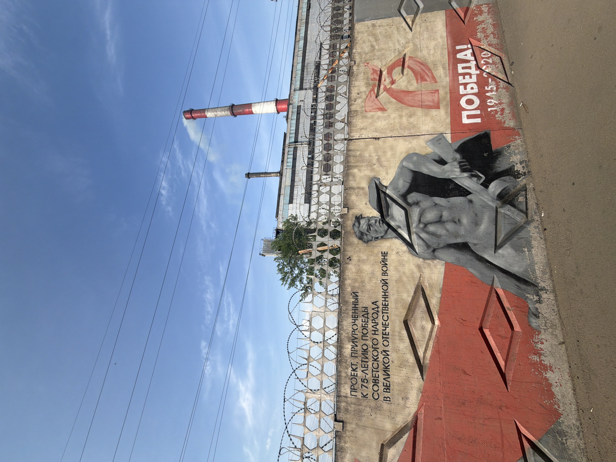 The image shows an outdoor scene in Birobidzhan, Russia. A section of a concrete wall is visible, painted with a mural commemorating the 75th anniversary of the Soviet people's victory in the Great Patriotic War (World War II). The mural features a stylized image of a muscular figure. The word "Победа!" (Victory!) and the years "1945-2020" are also prominently displayed on the wall. To the left of the wall, a tall industrial chimney with red and white stripes can be seen. A barbed wire fence is strung along the top of the wall and a larger structure behind it, adding a layer of security. Power lines stretch across the clear blue sky in the background. The image is taken from a low angle.