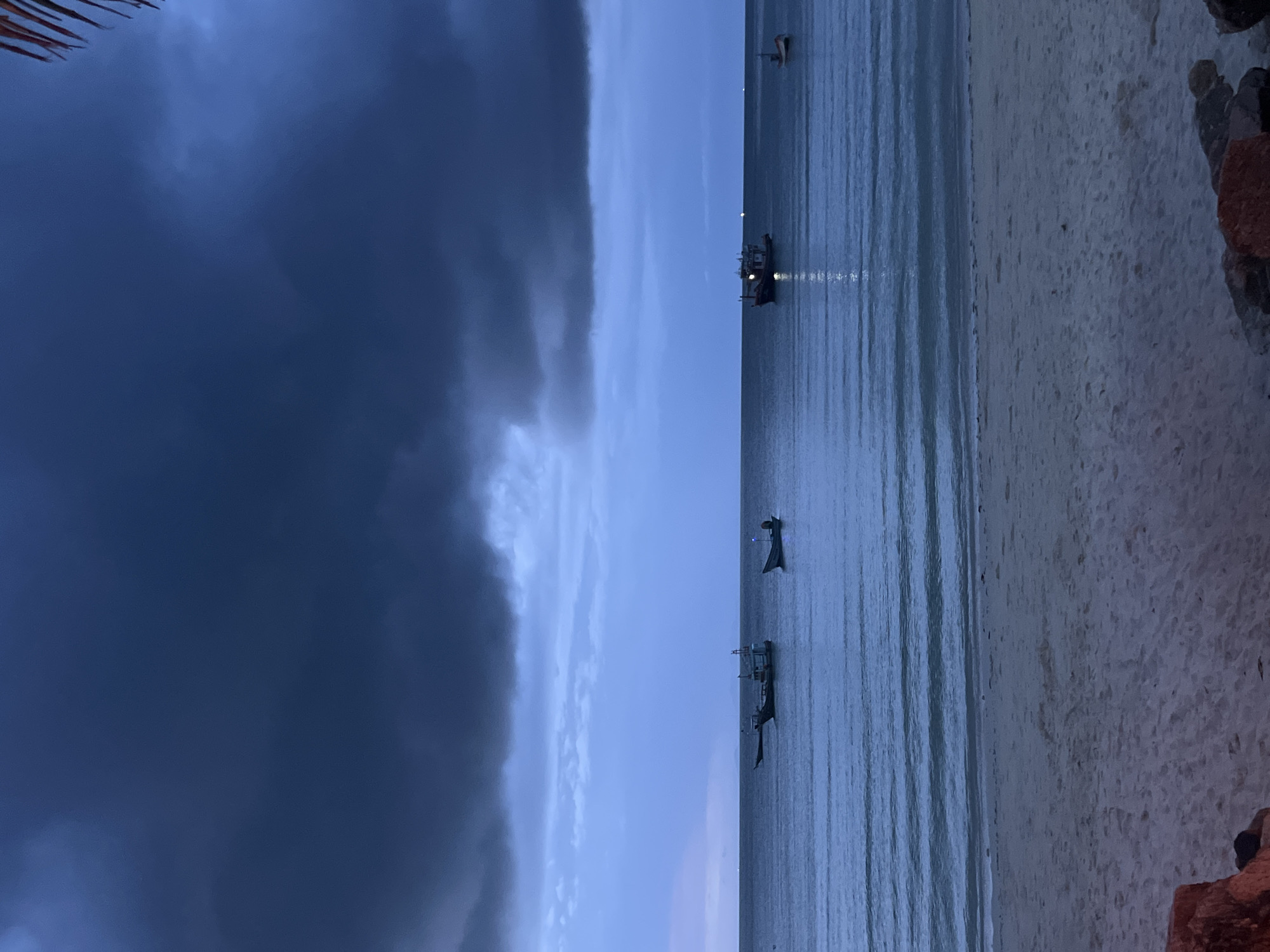 The image depicts a beach scene at dusk in Ko Pha-ngan District, Thailand. The sky is filled with dark, ominous clouds, suggesting an approaching storm. The sea is calm, with gentle waves lapping at the sandy shore. Several fishing boats are visible in the water, some illuminated by lights. The beach is sparsely populated, with only a few rocks visible in the foreground. The overall atmosphere is serene yet somewhat somber, reflecting the changing weather conditions.