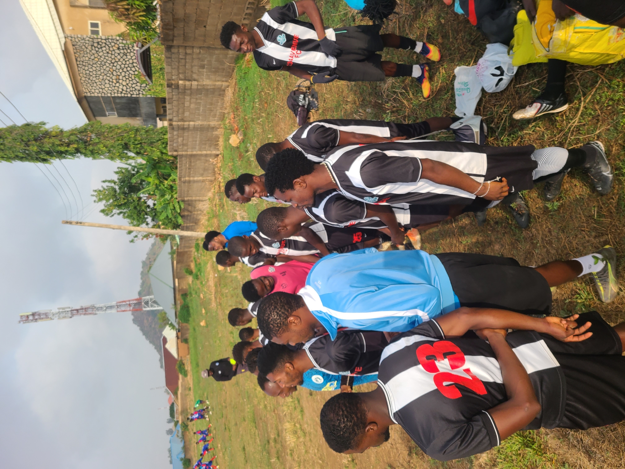 A group of male soccer players, mostly dressed in black and white striped jerseys, are engaged in a training session or stretching routine on a grassy field. They are arranged in a row, with some lying on their backs with arms behind their heads. Some individuals wear different colored shirts such as pink and blue. The players appear to be African and seem to be part of a team or club.
The background features a communications tower, buildings with stone and brick facades, and lush greenery. The presence of the tower suggests this location could be within the city limits of Bukuru, Nigeria, a location inferred from visual aspects that are consistent with the area's infrastructure and environment.