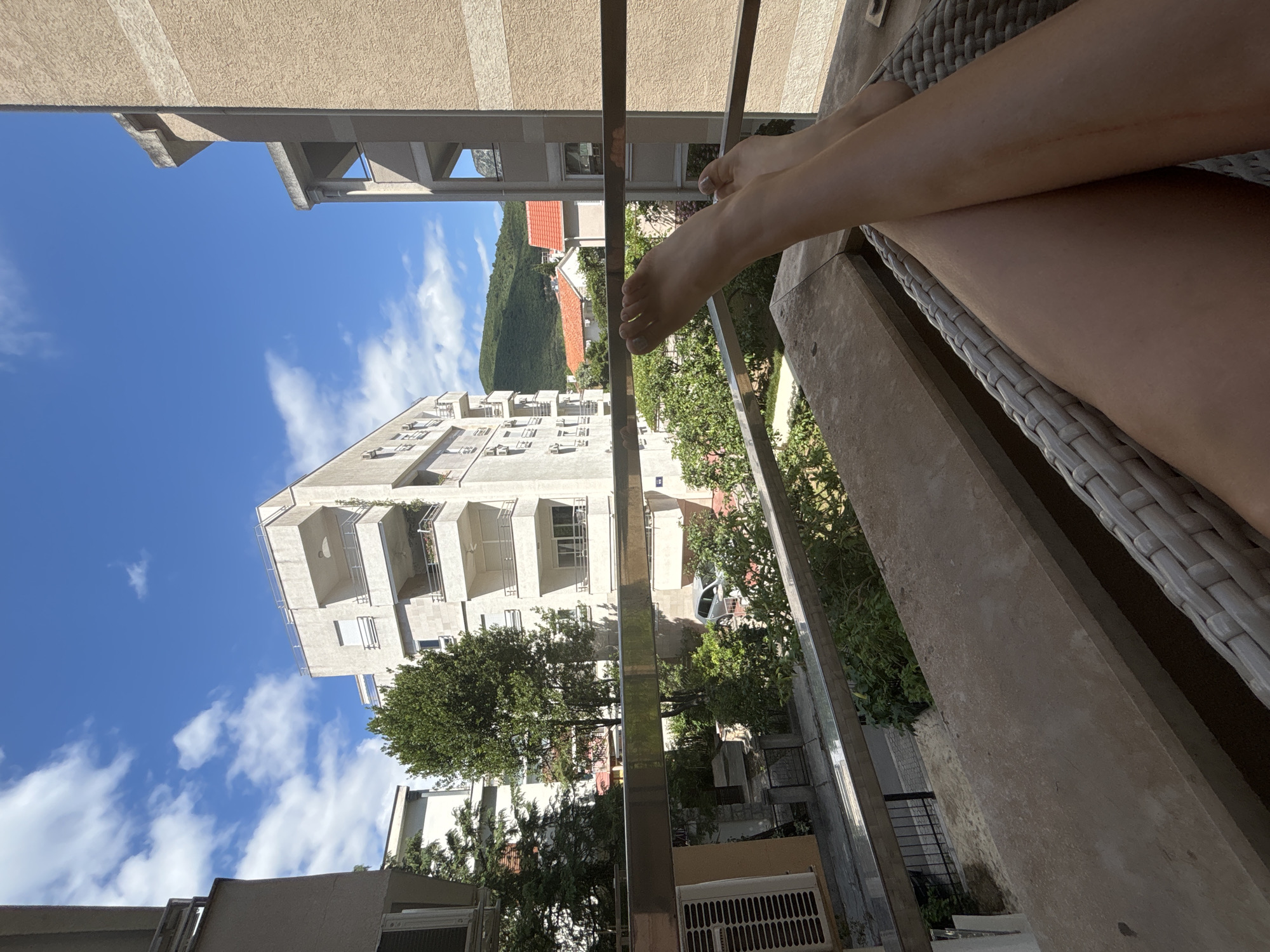 The image captures a view from a balcony in Budva, Montenegro, overlooking residential buildings and lush greenery under a bright blue sky dotted with white clouds. In the foreground, a person's legs and feet are resting on a woven chair, indicating a moment of relaxation. The view extends to modern apartment buildings with balconies and windows, a tree-lined street below, and a silver car parked. In the background, rolling green hills are visible, characteristic of the Montenegrin landscape. The scene suggests a warm, sunny day in a pleasant urban environment.