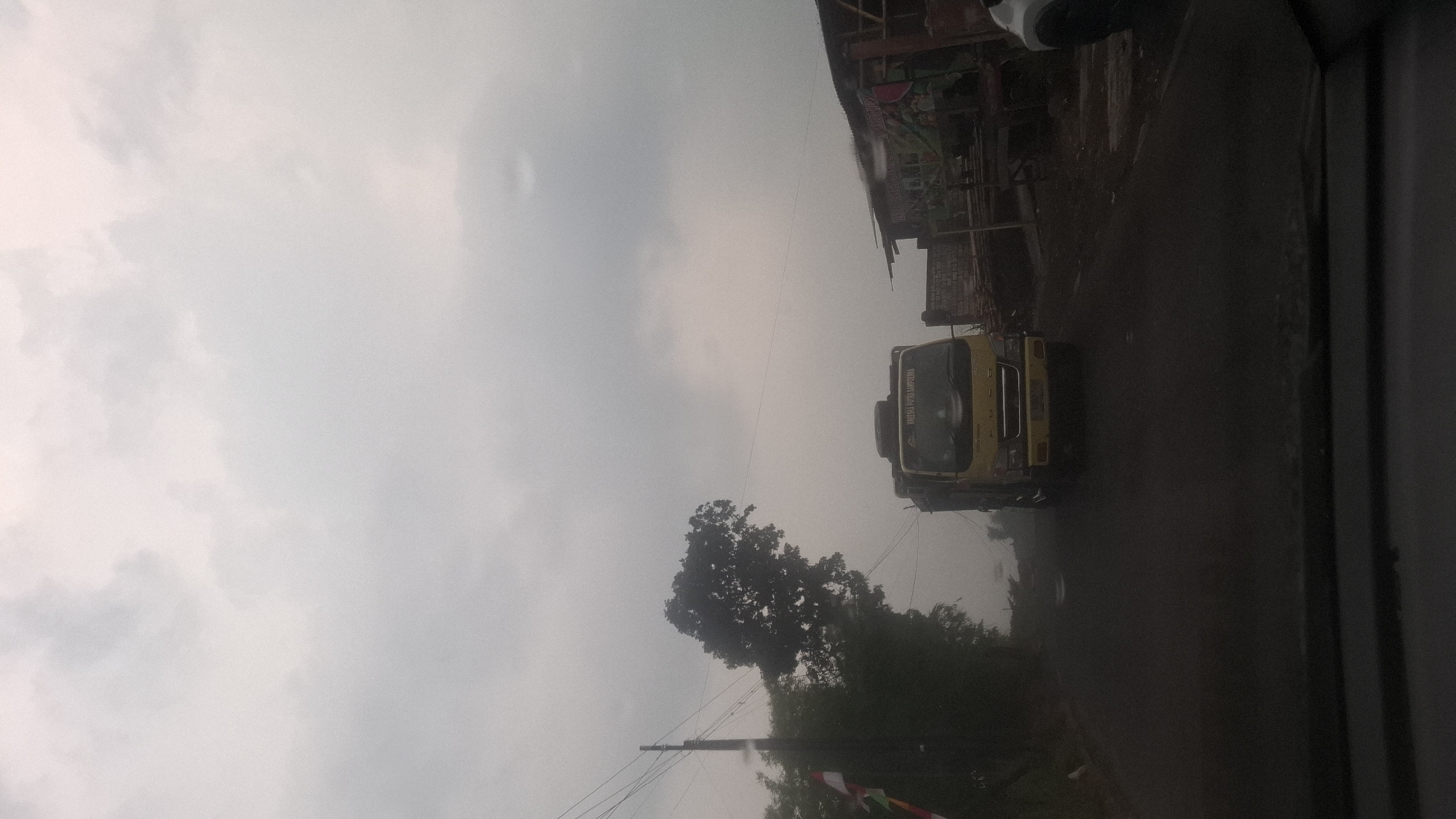The image shows a view from inside a vehicle looking out at a road. A yellow truck is driving down the road. To the left of the road, there are trees and a utility pole with wires. There is also a flag attached to a pole. To the right of the road, there are some buildings and what appear to be construction materials. The sky is cloudy. The scene appears to be in a rural or semi-rural area. The inscription on the truck reads "KRESNA PUTRA SAMPURNA".