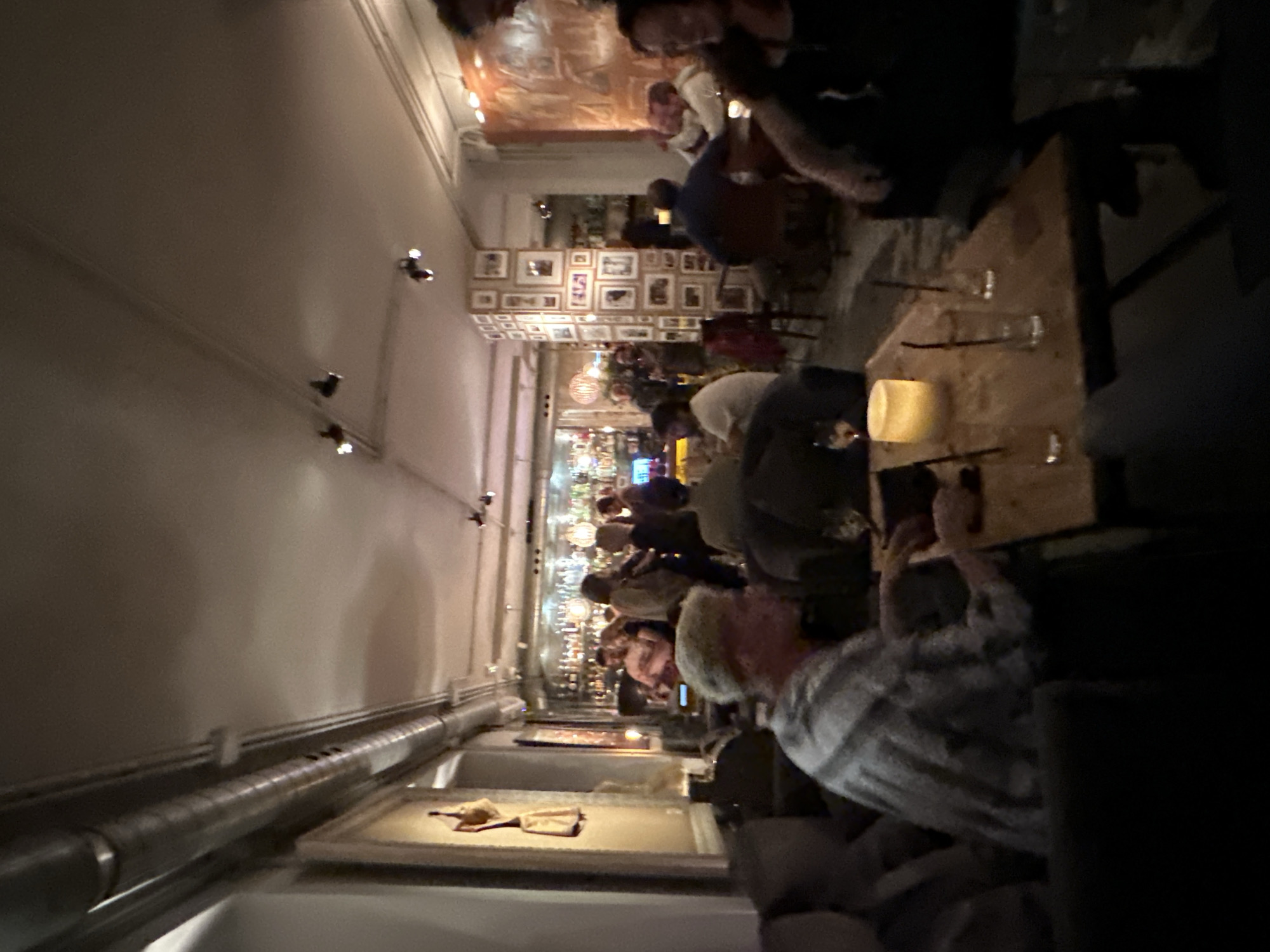 The image depicts a dimly lit, crowded interior of a bar or restaurant, possibly in Madrid, Spain. The scene features numerous people, many seated at a long wooden bar or table. Some individuals are engaged in conversation or looking at their mobile phones. A lit candle provides a focal point on the bar. The background reveals a wall covered in framed photographs or artwork. The overall atmosphere suggests a lively, social setting.