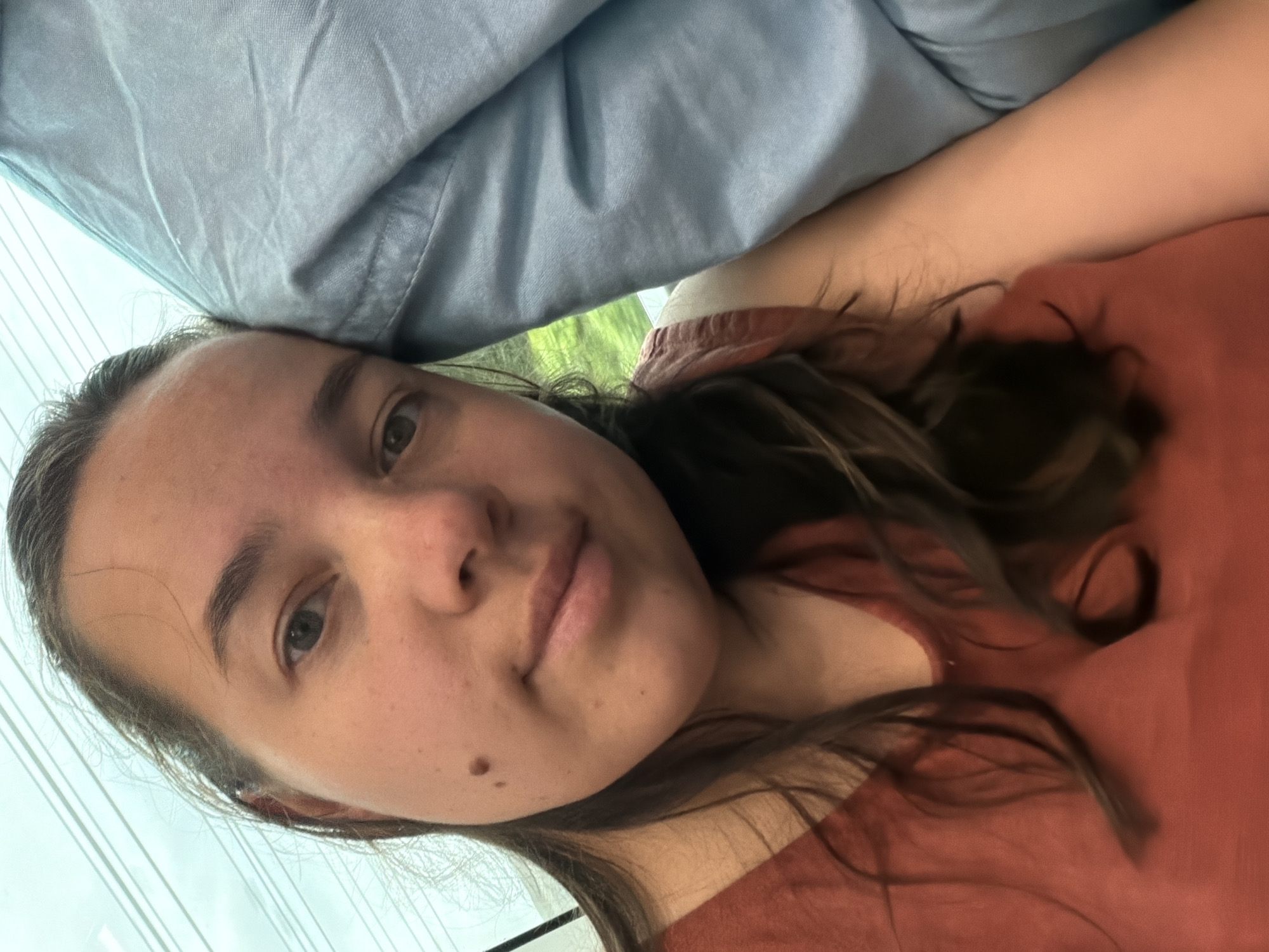 A young woman with long, dark hair is leaning her head on a light blue fabric, possibly a pillow or blanket. She is wearing a rust-colored top. Her face is turned towards the camera, and she has a mole on her left cheek. The background appears to be a window with a view of greenery, suggesting an outdoor or travel setting. The lighting is soft and natural. There are no discernible elements that would specifically place the scene in Yokohama, Japan.