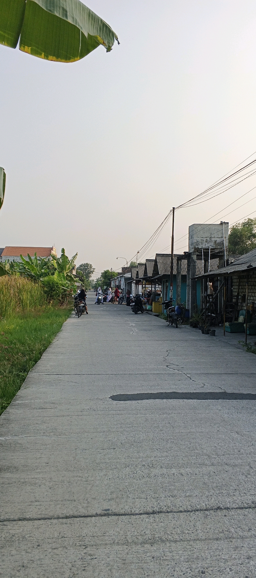 The image depicts a street scene in Latukan, Indonesia, likely in a rural or semi-urban area, characterized by a paved road, a line of small shops or stalls on the right, and vegetation on the left. The sky is overcast, suggesting early morning or late afternoon.
On the right side of the road, a row of buildings with corrugated metal roofs are visible. These appear to be small commercial establishments, possibly selling goods or offering services. Some have open fronts, revealing their interiors with shelves and items. Several motorcycles are parked in front of these shops. In the background, a concrete water tank stands on a raised platform, supported by pillars. A utility pole with numerous electrical wires is also prominent.
The left side of the road features lush green vegetation, including banana trees and tall grass. A building with a red tiled roof is partially visible behind the vegetation.
Several individuals are present on the road. A person is riding a motorcycle in the foreground, traveling away from the viewer. Further down the road, a group of people, some on motorcycles, are gathered. At least two individuals appear to be women wearing headscarves. One motorcycle has a passenger behind the rider, and another is being ridden by a person with a red bag. The overall atmosphere suggests a quiet, everyday moment in this Indonesian community. The road itself is concrete, with visible cracks and a dark, wet-looking patch indicating a possible spill or recent rain.