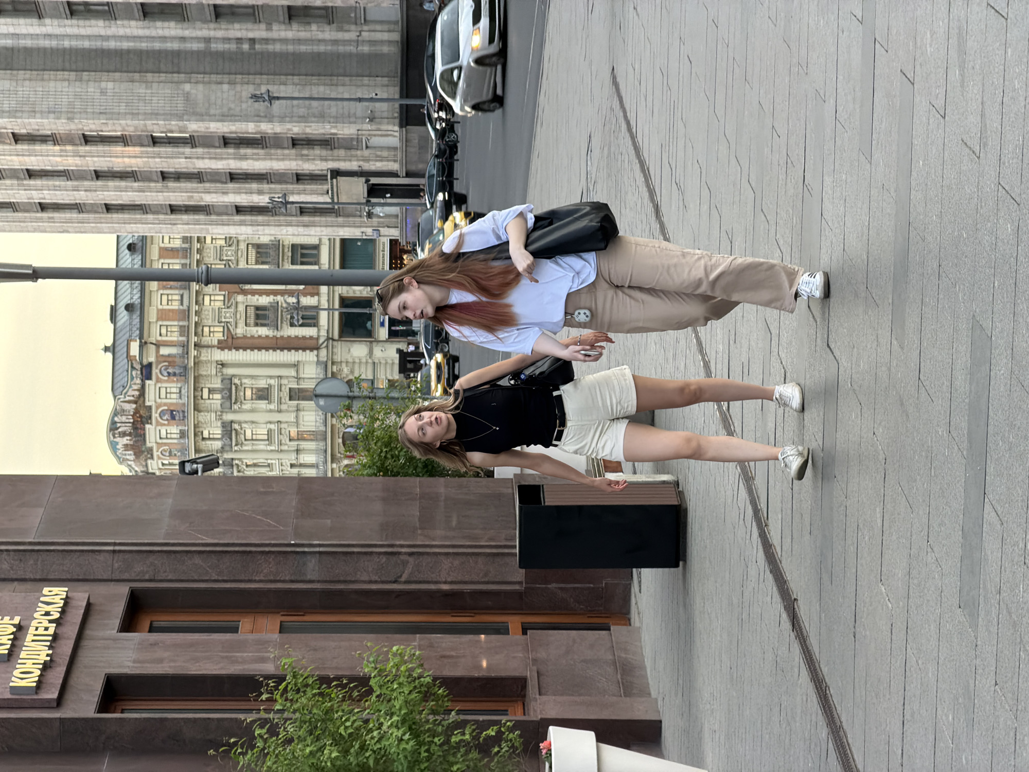 Two young women are standing on a sidewalk in Moscow, Russia. The woman in the foreground is wearing a black tank top and white shorts. The woman behind her is wearing a light blue shirt and khaki pants. A black shoulder bag hangs from the woman in the background. The sidewalk is gray and patterned. In the background are tall buildings and street traffic. A sign for "CAFE KONDITERSKAYA" is visible on a nearby building.