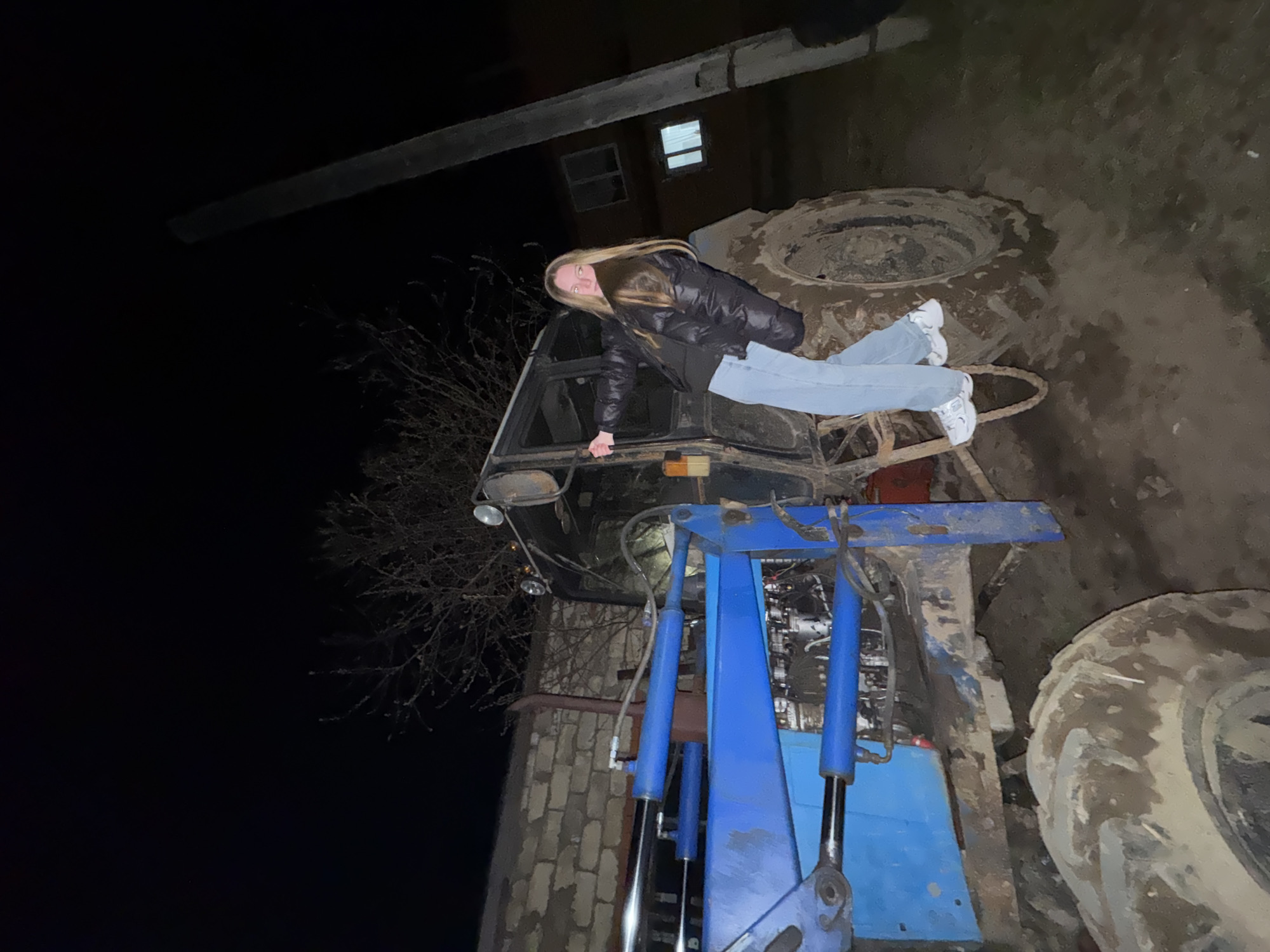 A nighttime scene depicts a woman reclining on a tractor or similar large vehicle. The tractor is dark in color and has large, muddy tires. It also features a blue hydraulic lift system. The woman is wearing a black puffy jacket, light blue jeans, and white sneakers. The background includes a building with a lit window, a bare tree, and a dark sky. The location is Zhigalki, Russia.