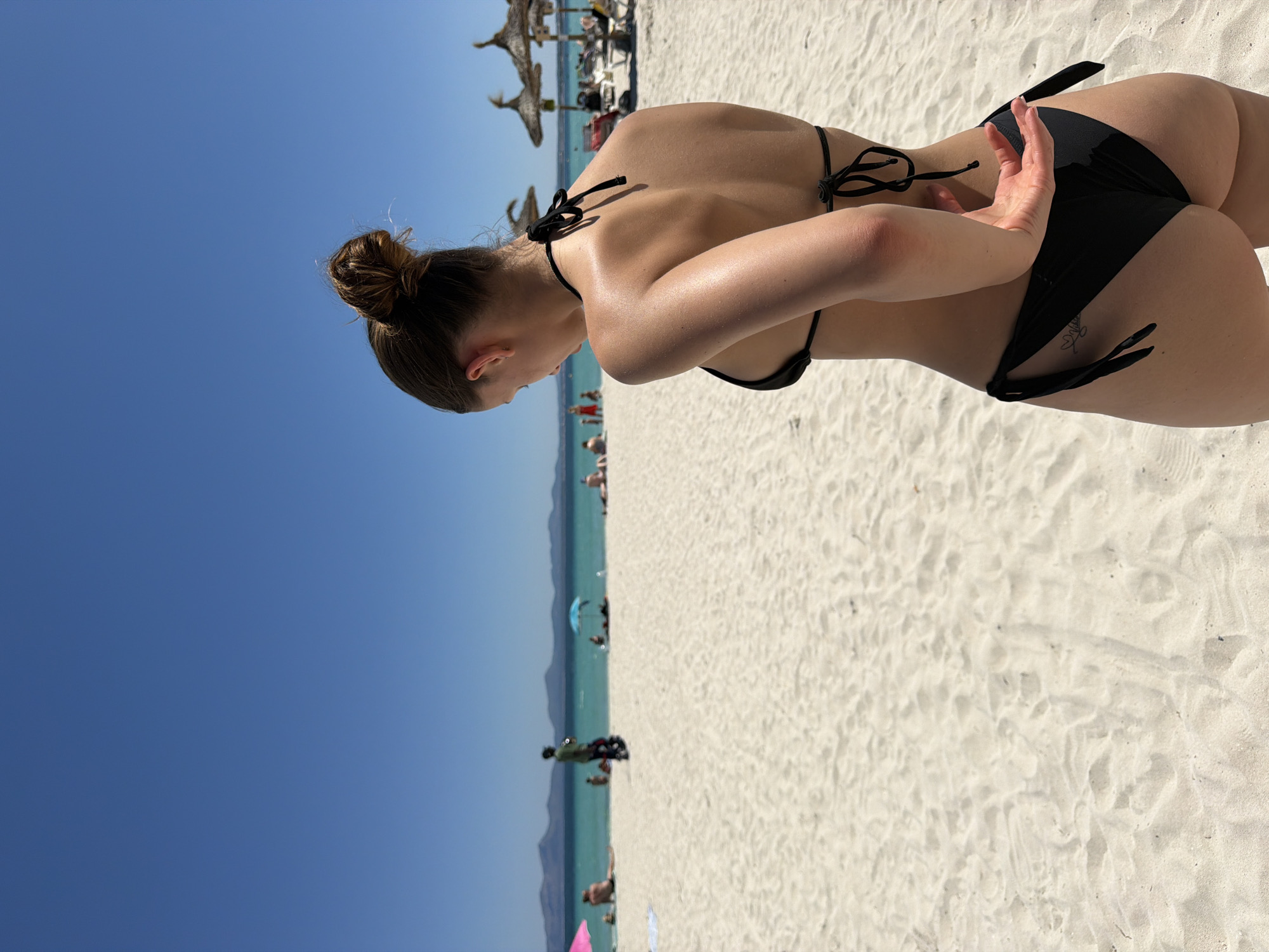A woman in a black bikini stands on a white sand beach on a sunny day. Her hair is styled in a bun, and she has her arm on her hip. In the background, the sea is turquoise, and there are people swimming and sunbathing. Umbrellas and some palm trees are visible in the distance. The sky is a clear blue. The location is Alcúdia, Spain.