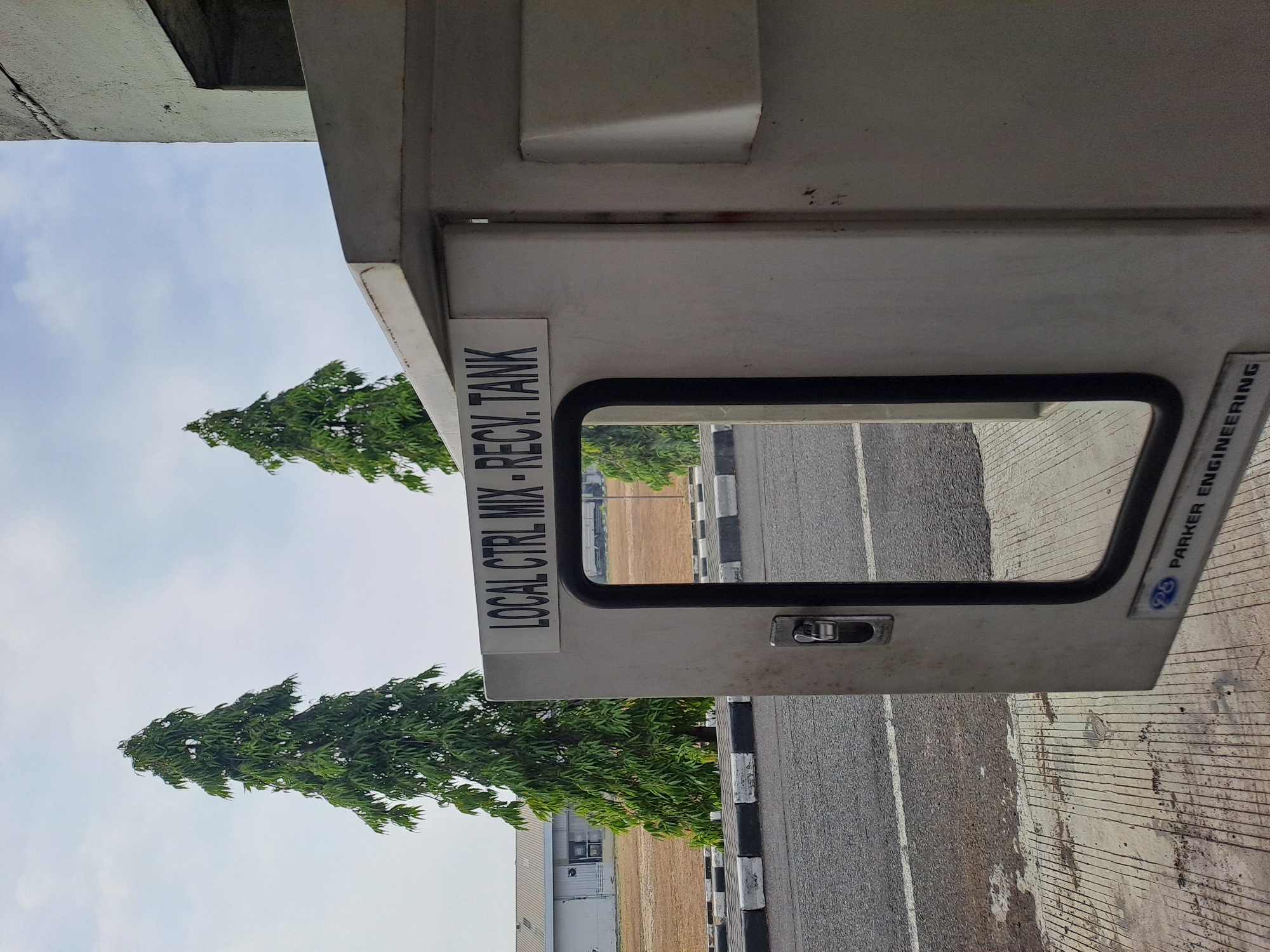 The image shows an outdoor scene in Purwakarta, Indonesia, with a gray utility box in the foreground. The box has an open door revealing a mirrored surface and a small latch. A sign on the box reads "LOCAL CTRL MIX - RECV. TANK". To the left of the box is a paved road with white and black striped curbs, and beyond that, a grassy area with a large industrial building in the background. Two tall, green trees stand between the road and the building. The sky is partly cloudy. A logo with the text "PARKER ENGINEERING" is visible on the bottom of the utility box. The scene appears to be calm and static, with no people or animals present.