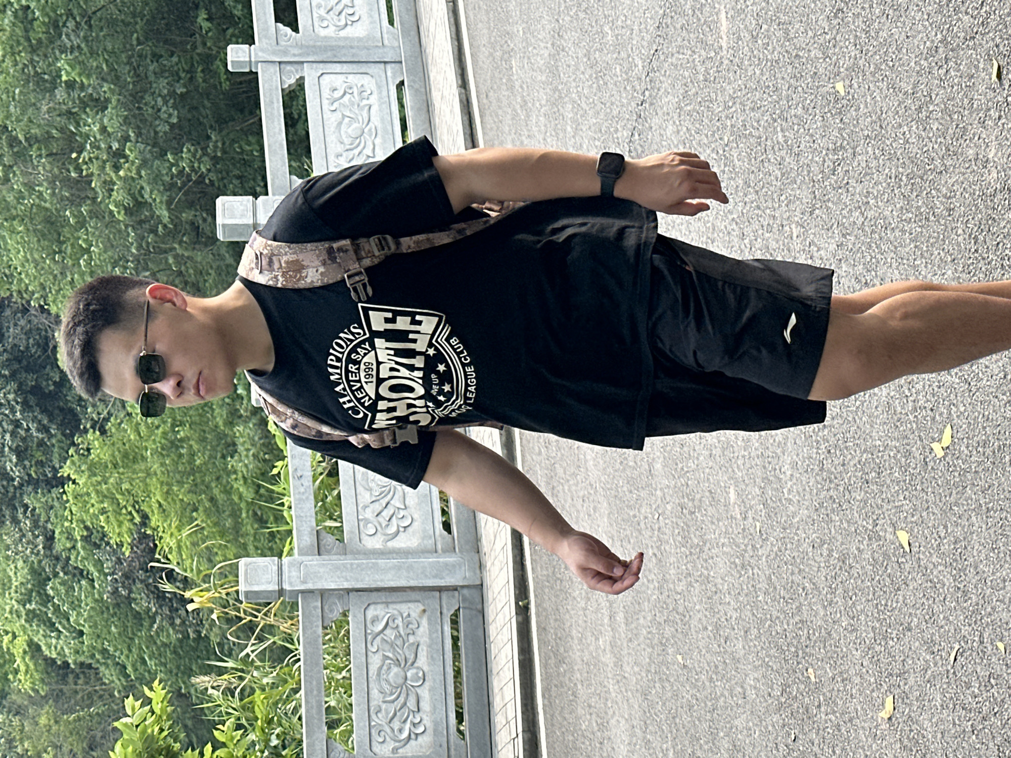 A young man stands outdoors on a sunny day in Liangjiang, China. He is positioned facing slightly to his right, looking away from the camera. He appears to be of East Asian descent, with short, dark hair and sunglasses. He is dressed casually in a black short-sleeved t-shirt, black athletic shorts with a white logo on the left leg, and a black digital watch on his left wrist. A camouflage-patterned sling bag or cross-body bag is visible over his right shoulder.
The t-shirt features prominent white text, which includes "CHAMPIONS NEVER SAY 1999" and a large word that appears to be "CHIPOTLE" or a similar stylized name, with "WE UP LEAGUE CLUB" written below it.
The setting suggests a park or scenic area, possibly near a body of water or elevated pathway. Behind the man, to his left, is a dense background of lush green trees and foliage, indicating a natural environment. To his right and in the middle ground, there is a grey stone railing or balustrade with ornate, carved designs. The ground he stands on is a grey, textured paved surface, possibly asphalt or concrete, with small light-colored flakes or debris scattered across it. The bright, even lighting indicates it is daytime with clear weather. The man's expression is neutral, and he appears to be in a relaxed pose, either standing still or mid-walk.