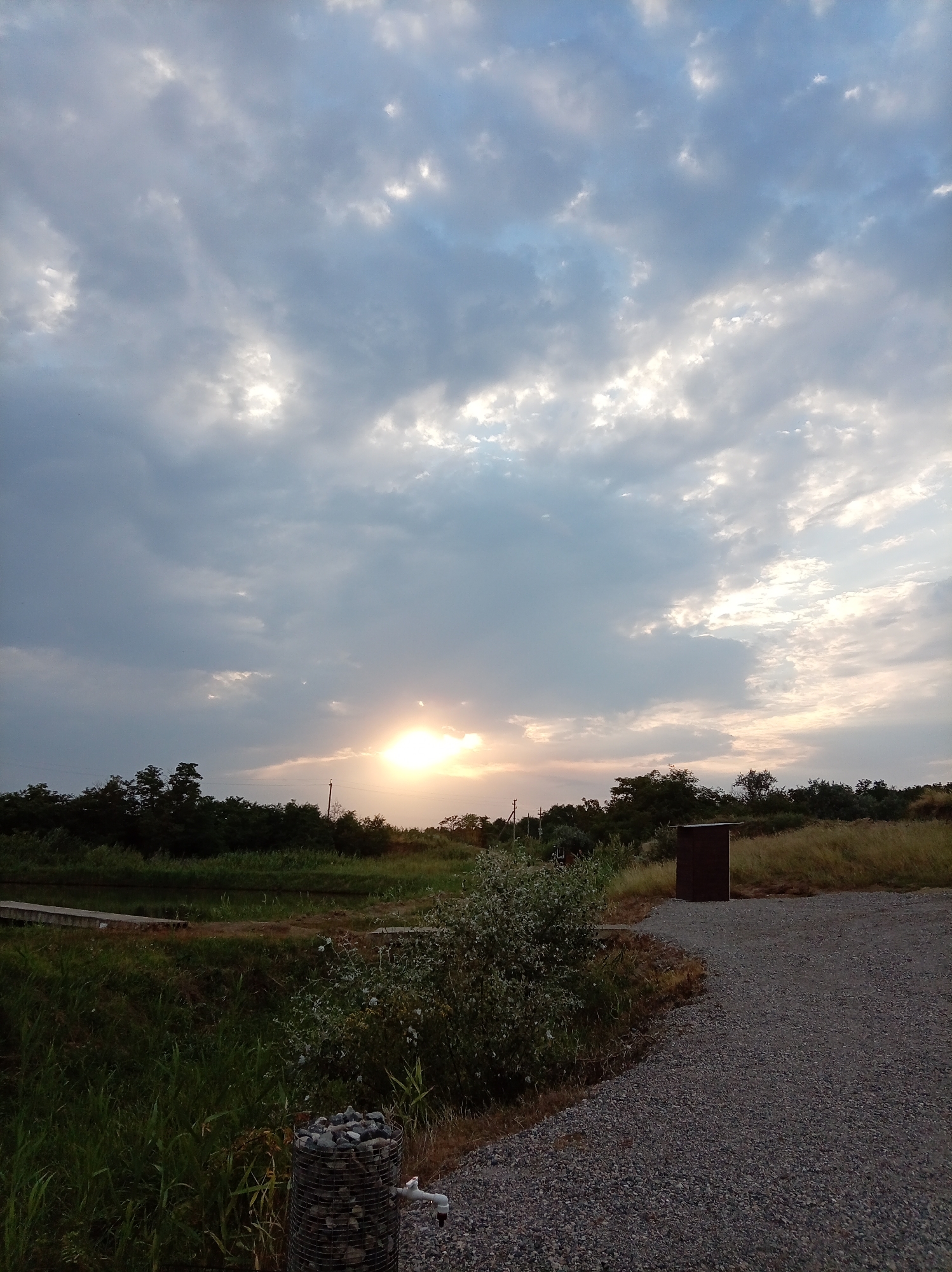 The image depicts an outdoor scene with a cloudy sky at sunset. The sun is visible as a bright orb peeking through the clouds, casting a warm glow. In the foreground, a gravel path leads towards the right side of the frame. To the left of the path, there is tall green grass and some vegetation. A cylindrical structure filled with stones and a faucet is visible in the lower left corner. Beyond the grass, there is a body of water, possibly a pond or canal, with a wooden walkway. A dense line of dark green trees borders the far side of the water. In the distance, more trees and dry grass are visible under the muted sky. A small, dark brown, shed-like structure stands in the middle ground. The overall atmosphere suggests a rural or semi-rural setting, possibly a park or an area near a body of water, in or near the city of Grazhdanskoye, Russia. There are no people or animals visible in the frame.
