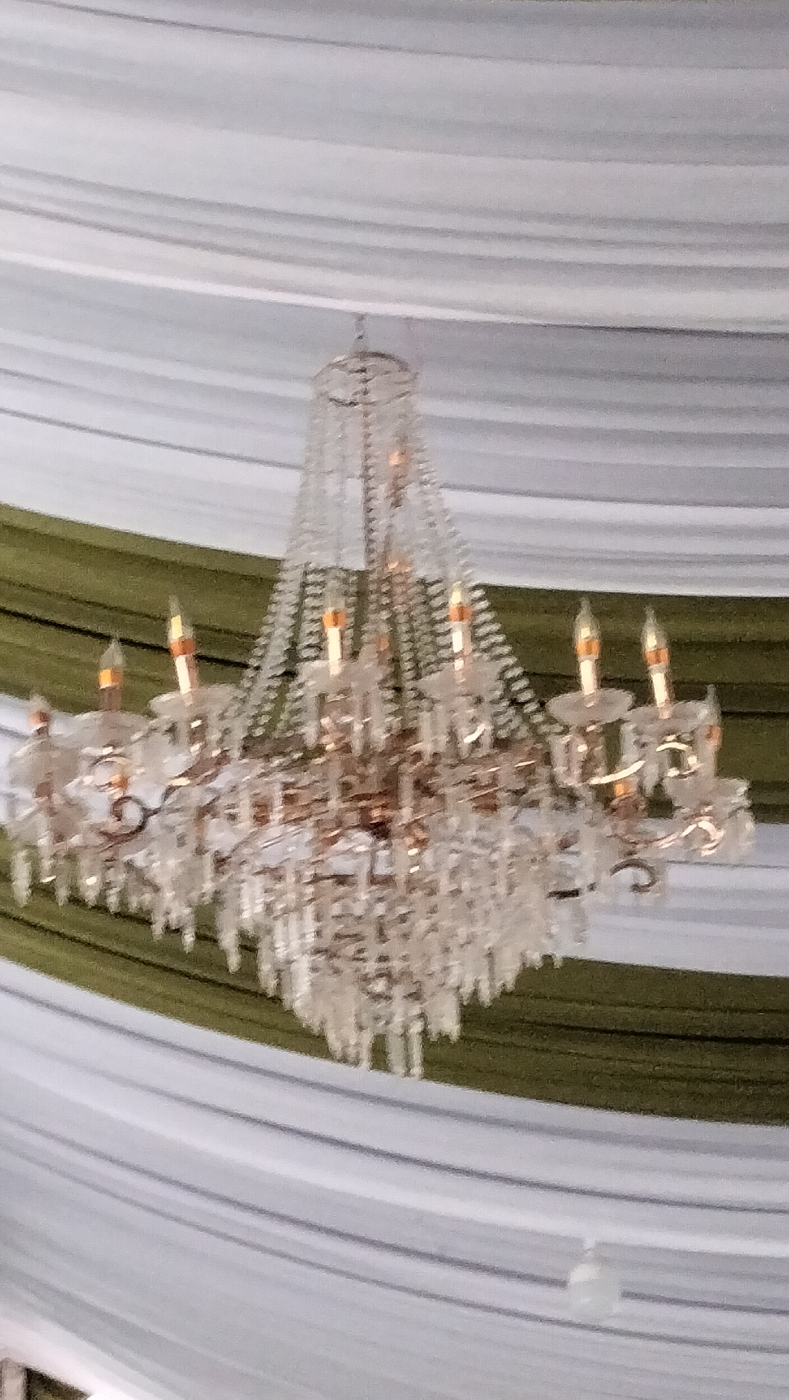 A chandelier hangs from a draped ceiling made of white and green fabric. The chandelier is ornate, made of clear glass or crystal, with multiple arms holding faux candles. The fabric ceiling appears to be part of a temporary structure, possibly a tent or a banquet hall. The overall scene suggests an event or celebration. The image does not contain information about the city of Lhokseumawe, Indonesia.