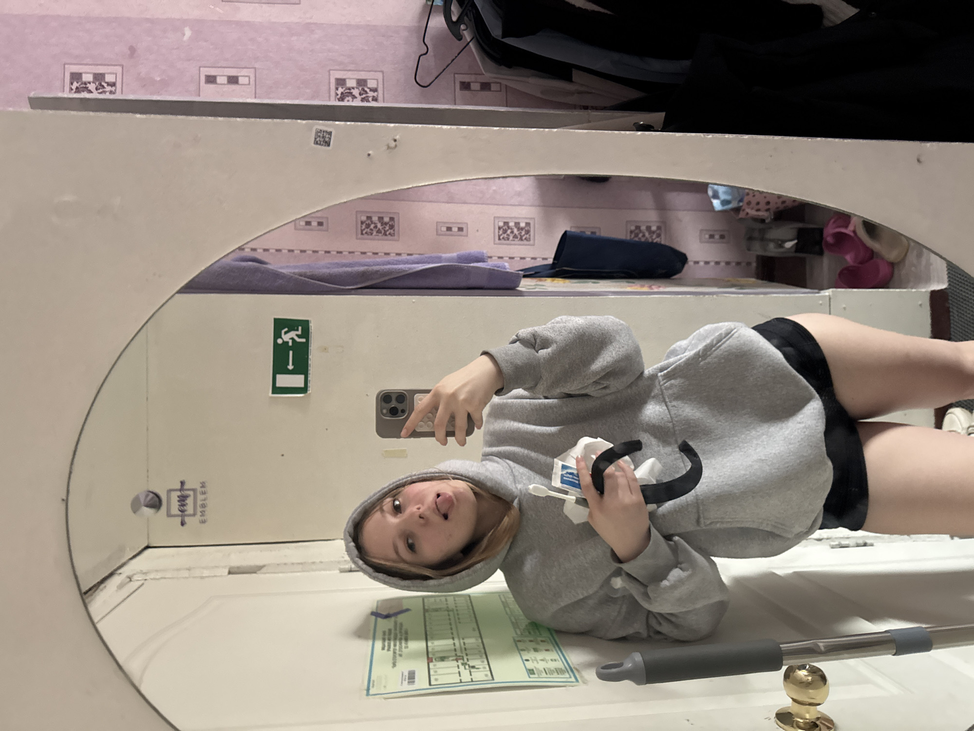 A woman is reflected in a large, round mirror. She holds a smartphone in one hand and a toothbrush and a piece of tissue in the other. She is wearing a gray hooded sweatshirt and black shorts. The mirror is mounted on a white door, on which an evacuation plan is posted. A mop leans against the door. Above the door are shelves, upon which are toiletries and clothing. The room is located in Perm, Russia, likely in a dorm or hostel, as evidenced by the emergency exit sign and simple furnishings.