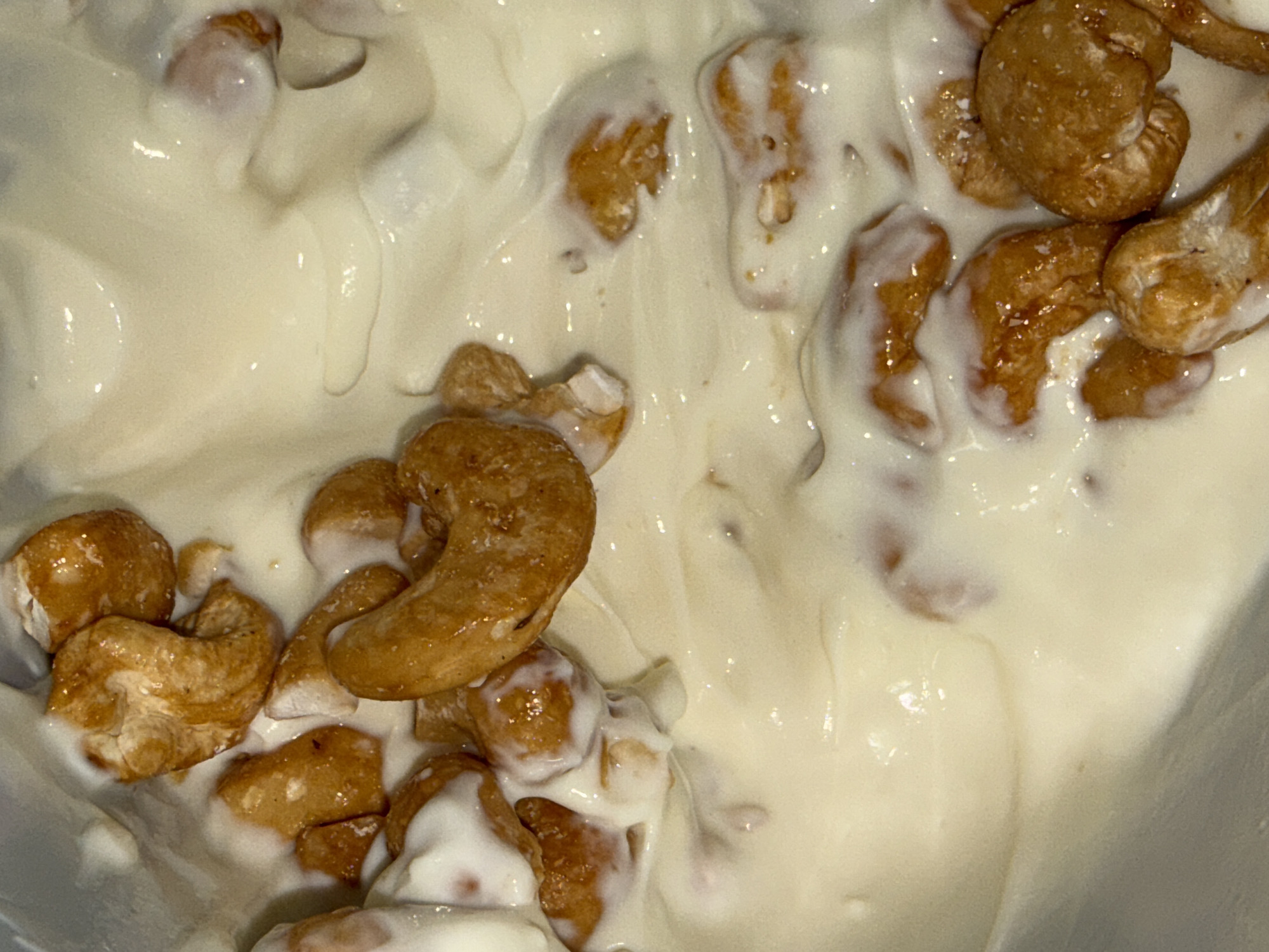 The image shows a close-up of cashews covered in a white, creamy substance, possibly yogurt or a similar dairy product. The cashews are scattered and partially submerged in the cream. The scene lacks any discernible contextual clues to place it specifically in Igboefon, Nigeria, or any information related to people, actions, or the environment.