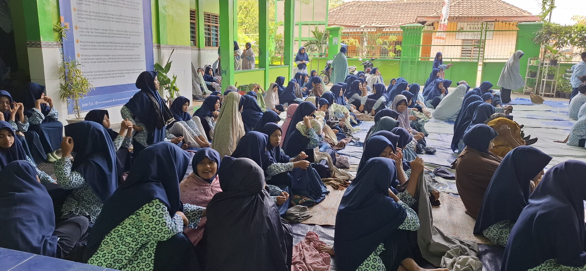 The image depicts a group of individuals, primarily young women and girls, seated on the ground in an outdoor covered area. They are wearing hijabs and modest clothing, many in shades of blue and white. The setting appears to be a school or community gathering space in Wonorejo, Indonesia, suggested by the architecture and vegetation. A green banner with text in Indonesian is visible on the left. Some individuals are looking towards the camera, while others are engaged in conversation or looking down. The ground is covered with mats and tarps. In the background, there are buildings with tiled roofs and a fence. The overall atmosphere seems to be one of a communal event or educational activity.
