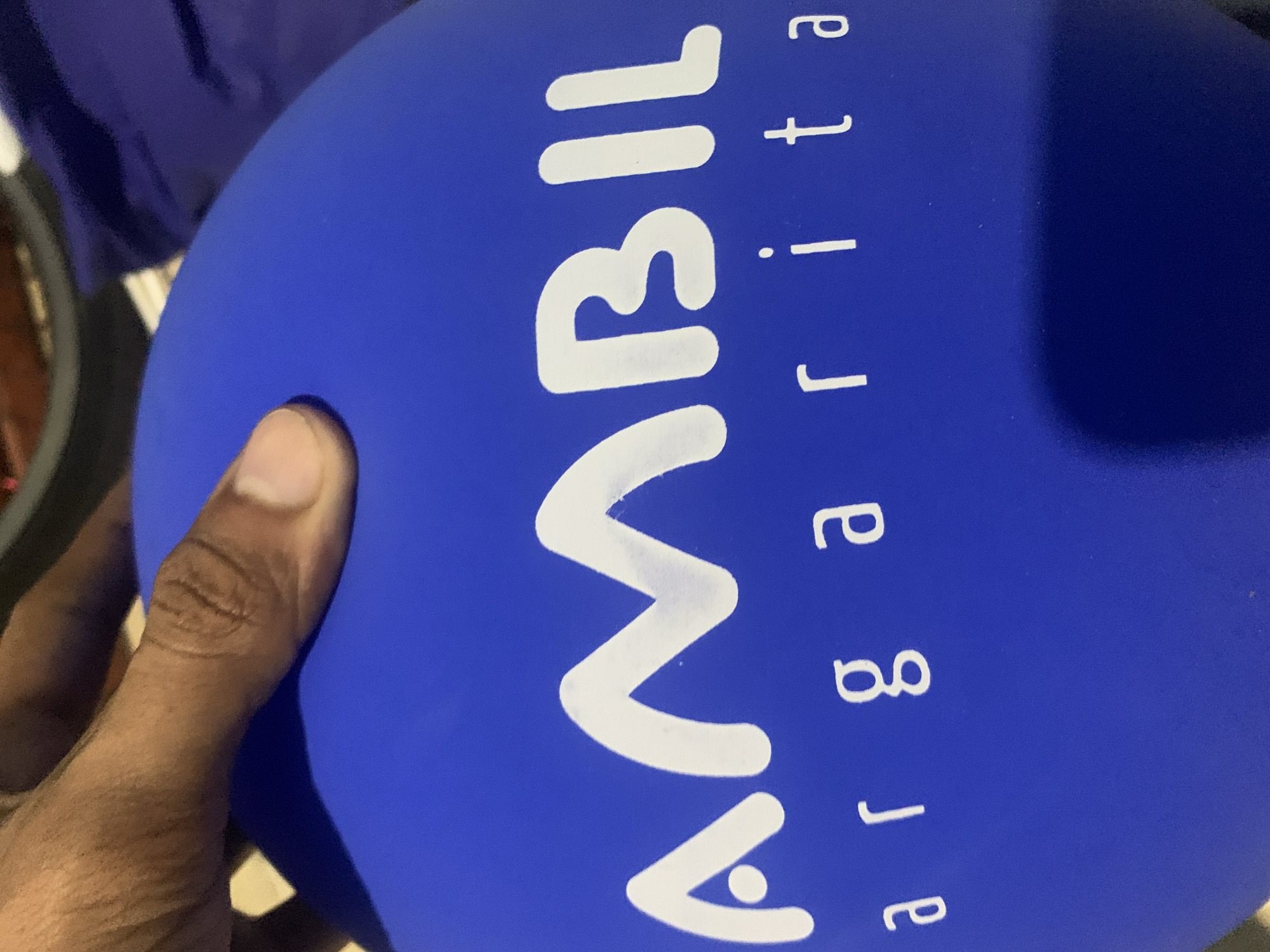 A person's hand, with a dark skin tone, is holding a blue inflatable balloon. The scene appears to be indoors, with blurred, dark garments and indistinct objects visible in the background. The activity involves someone holding the balloon, possibly at an event or gathering. Notable details include the solid blue color of the balloon and the clear skin texture of the hand. The image does not provide visual cues for the specified location of Santa Ana, Venezuela, nor for the time of day or emotions.
White text is prominently displayed on the balloon. The main, larger text reads "AMARIL", featuring a stylized first 'A' with a small dot inside, a wavy 'M', and an 'R' resembling a backward '3'. To the right of this word, smaller letters are arranged vertically. Reading from top to bottom, these letters clearly spell "a", "r", "j", "e", "t", "a", forming the word "arjeta". This text combination, "AMARIL arjeta", suggests a brand name "AMARIL" followed by a descriptor, possibly implying "Tarjeta" (Spanish for 'card'), with the initial 'T' either missing, stylized as 'a', or out of frame.