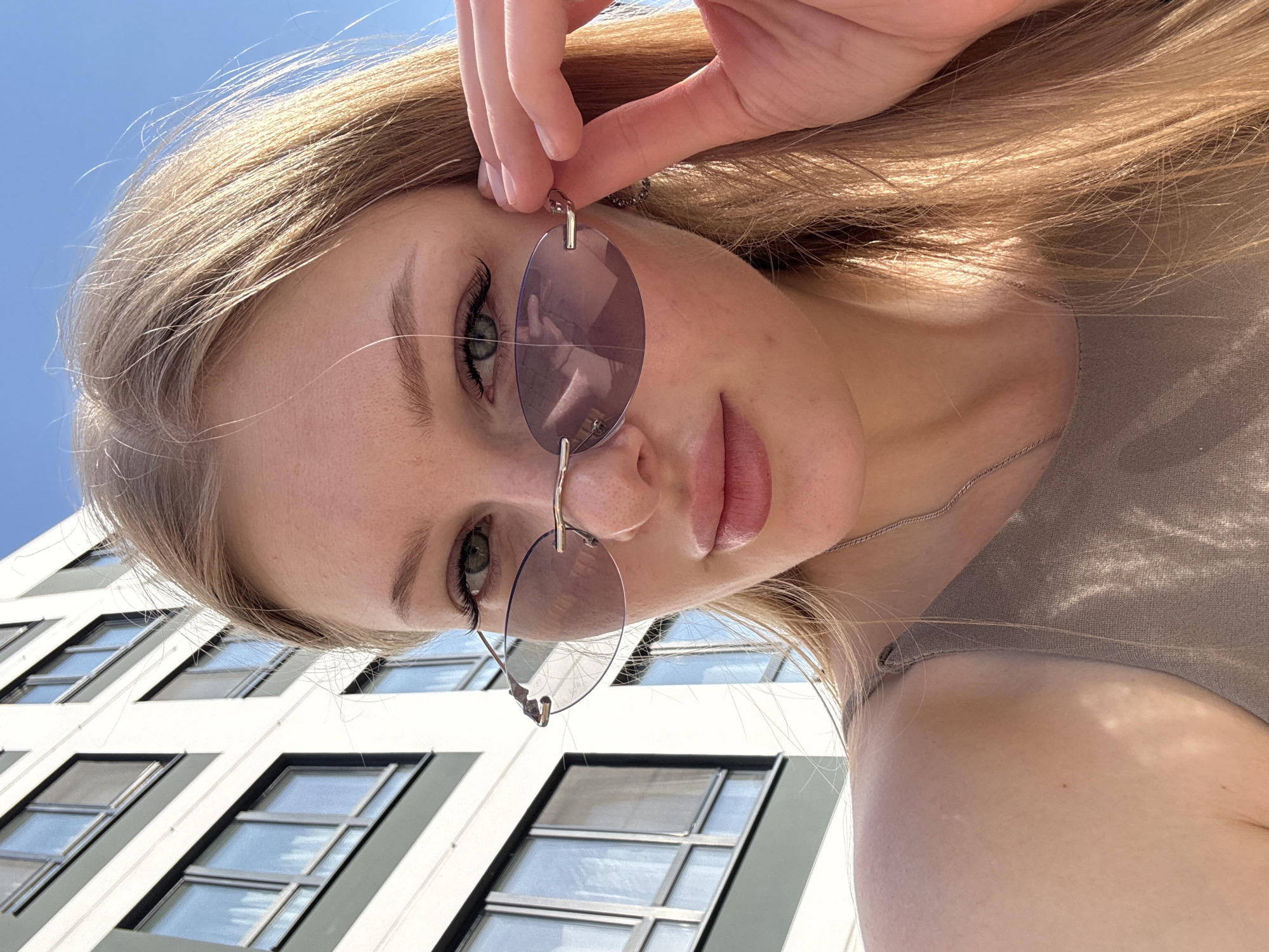 A young woman with blonde hair and green eyes is holding a pair of round, purple-tinted sunglasses. She is wearing a light-colored top and a thin silver necklace. The image is taken from a low angle, looking up at her face. In the background, there is a multi-story building with square windows. The location appears to be an urban area, possibly in Perm, Russia, based on the architectural style of the building.