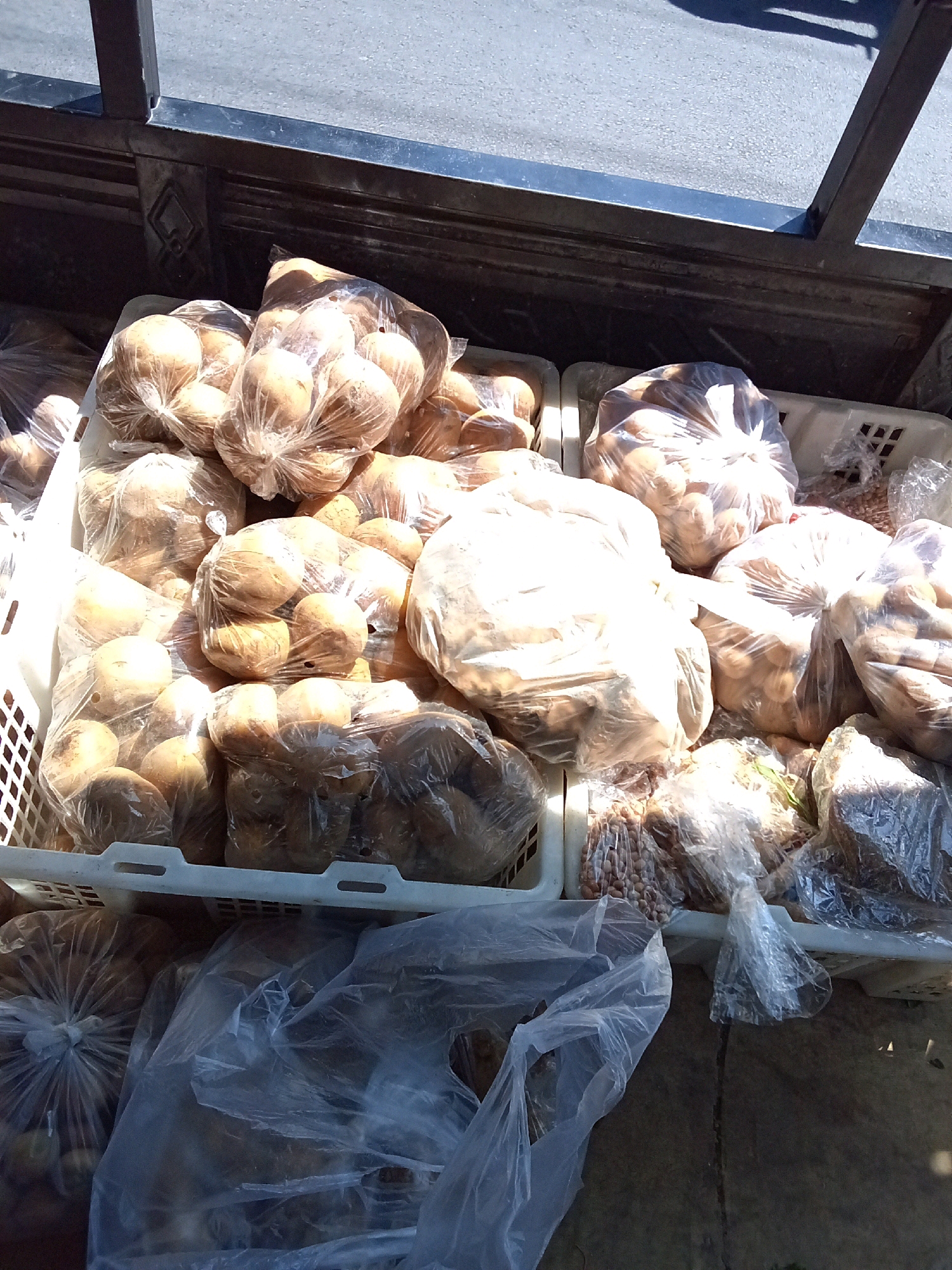 In Ngasem, Indonesia, a close-up shot reveals a market stall laden with fresh produce. The foreground is dominated by several white plastic crates overflowing with potatoes, each batch contained within clear plastic bags. The potatoes appear to be of varying sizes and are a light brown color. Some bags are tightly bundled, while others are more loosely packed, allowing the texture of the potatoes to be visible. The crates are arranged side-by-side on a surface that appears to be concrete or stone. Behind the crates, a dark metal railing is visible, casting a shadow on the asphalt or paved ground beyond, suggesting an outdoor market setting. The lighting indicates it is daytime, with sunlight creating bright highlights on the plastic bags and casting shadows. No people are visible in the frame, focusing the attention entirely on the abundance of potatoes.