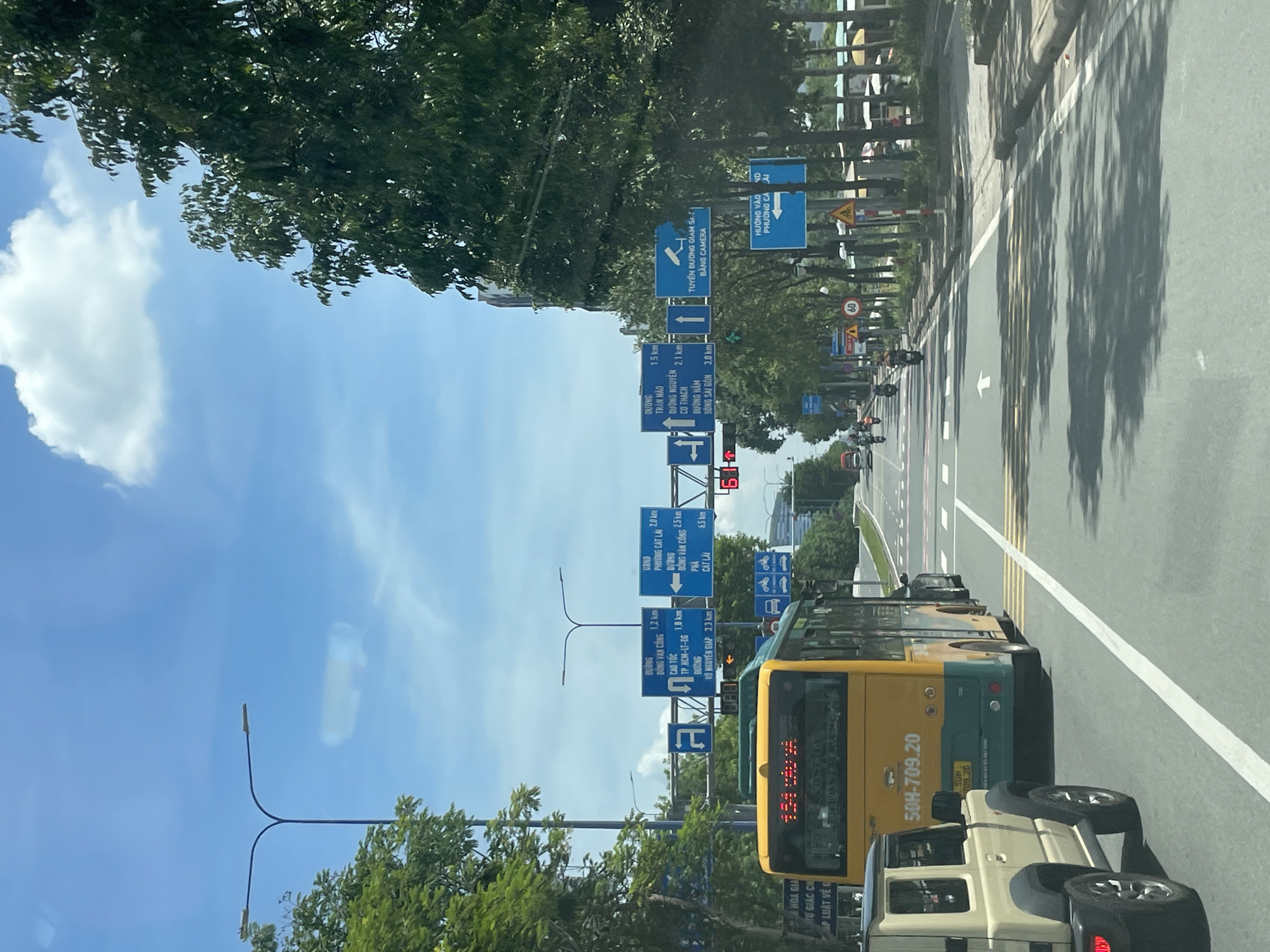 The image depicts a daytime view of a multi-lane road in Thủ Đức City, Vietnam. A yellow and green bus, identified by the license plate "50H-709.20," is in the foreground, occupying the right lane. To the left of the bus, a light-colored SUV is also visible. Traffic signs indicating directions and distances to various locations such as Cần Thơ, Cần Giờ, and Đồng Nai are present above the road. The road is marked with white lines and a pedestrian crossing. Trees line the sides of the road, and a bright blue sky with scattered white clouds is overhead. Several motorcycles are seen further down the road, indicating ongoing traffic.