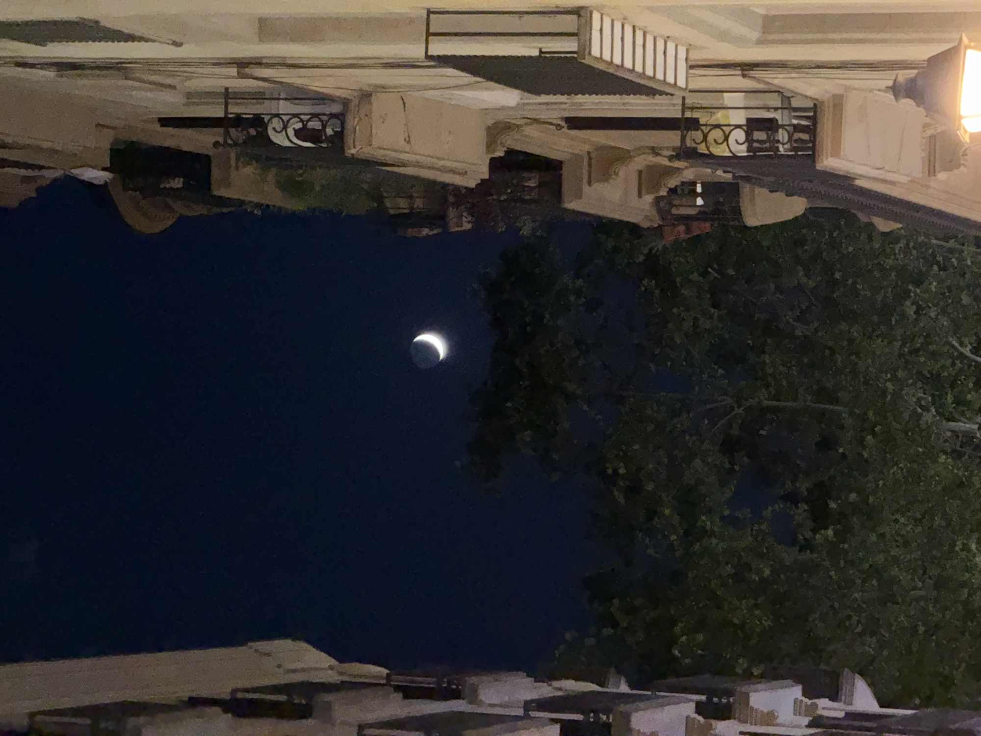 The image captures a crescent moon in a dark blue night sky, positioned above the canopy of lush green trees. The scene is framed by the facades of two buildings on either side, suggesting an urban setting. The building on the left is partially visible, with its textured wall in muted tones. The building on the right is more prominent, showcasing a cream-colored facade with balconies featuring ornate wrought-iron railings. One balcony displays chairs and a small table. A street lamp with a warm, yellow light illuminates the lower right portion of the frame. The composition creates a sense of depth, with the moon and trees appearing distant, while the buildings are closer to the viewer. The overall atmosphere is tranquil and nocturnal.