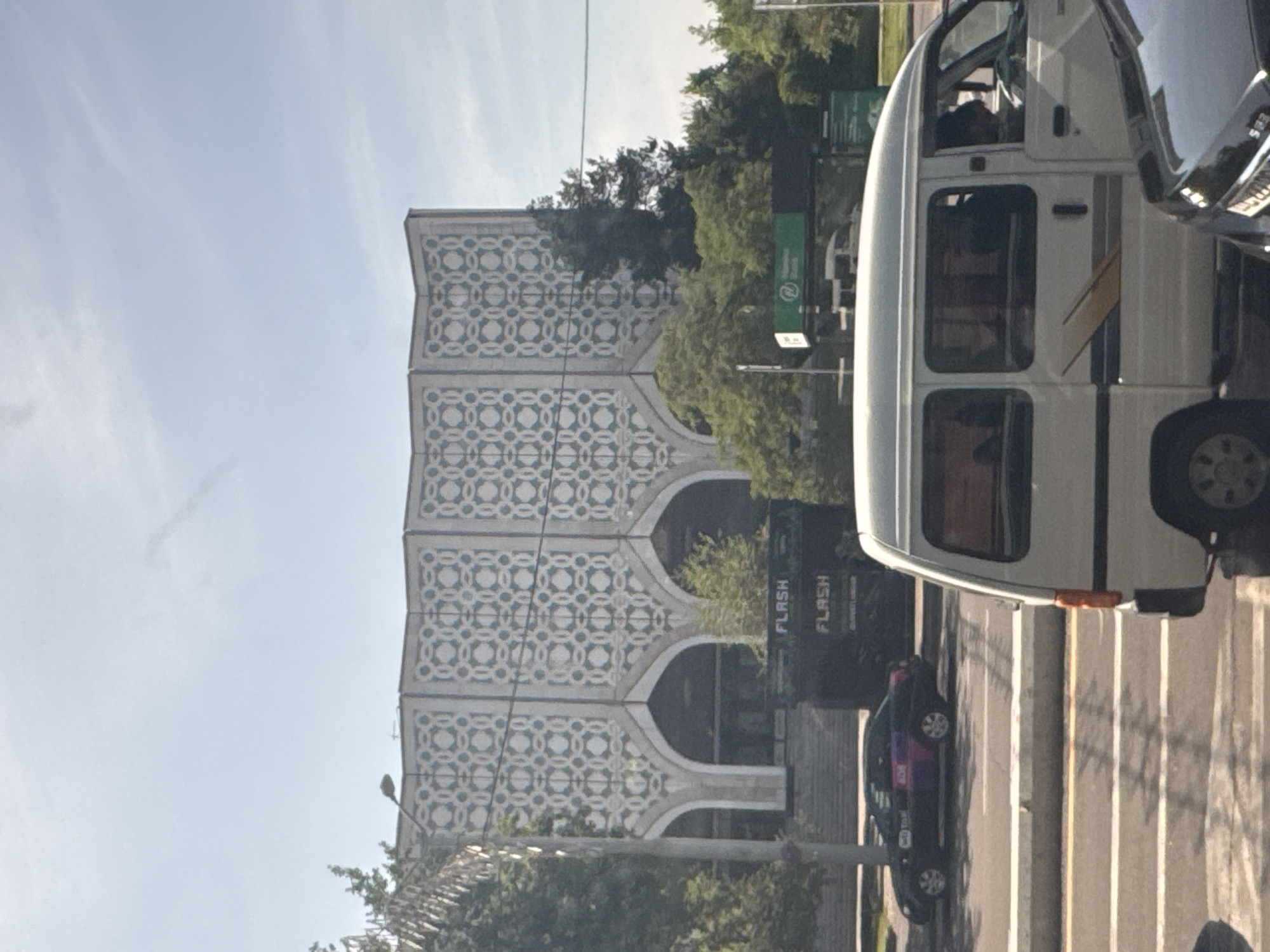 The image shows a building in Tashkent, Uzbekistan, characterized by its patterned facade with repeating circular and square motifs, as well as arched openings. In the foreground, a white van partially obscures the view of the street, which has multiple lanes with zebra crossing lines. A black car is also visible on the road. Trees and greenery flank the building, and signage is present, including "FLASH". The sky is clear with some wispy clouds. The photo appears to have been taken from a vehicle.